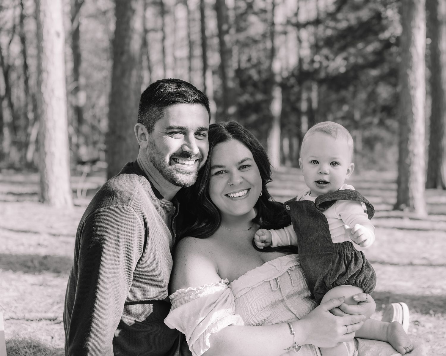 A black and white portrait of a family smiling together in a forest setting.