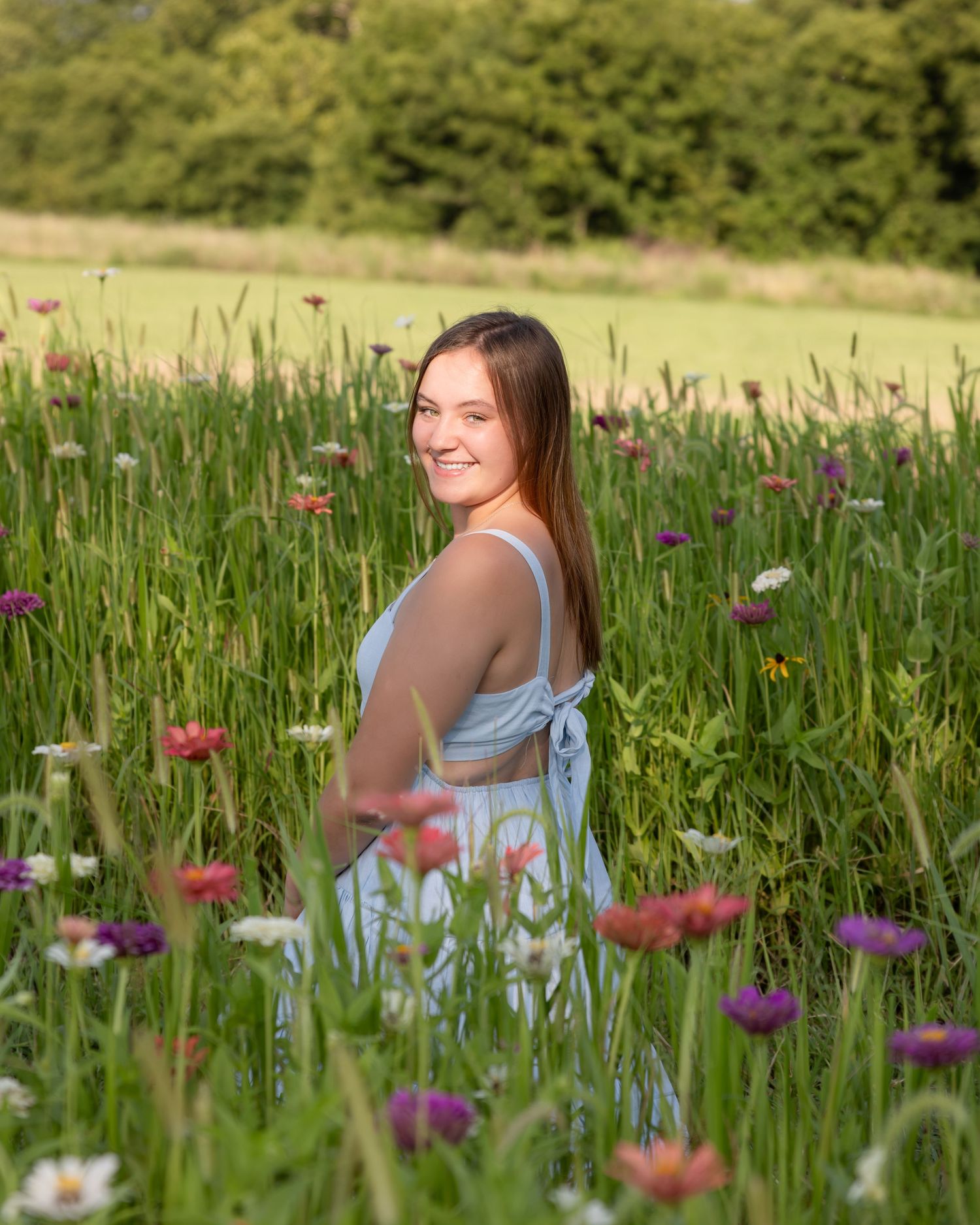 Young woman in light blue dress smiling among colorful wildflowers in a lush green field.