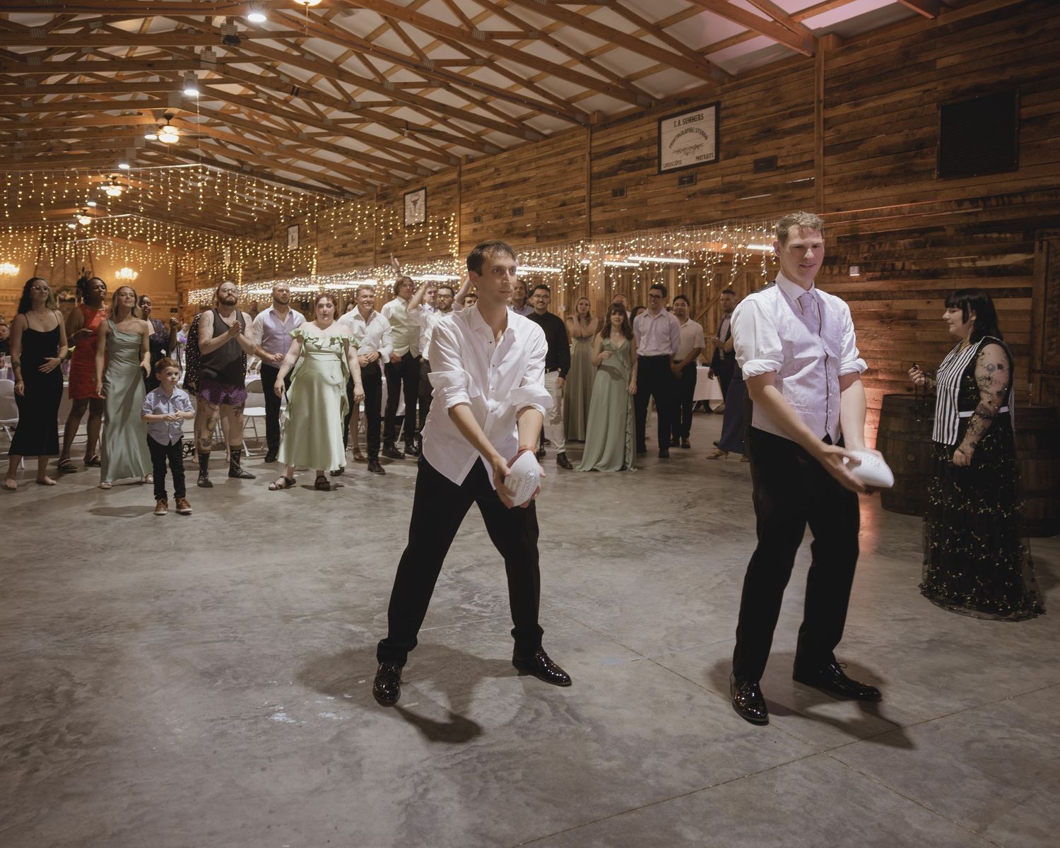 Two men performing a choreographed dance at a rustic barn wedding reception while guests watch.