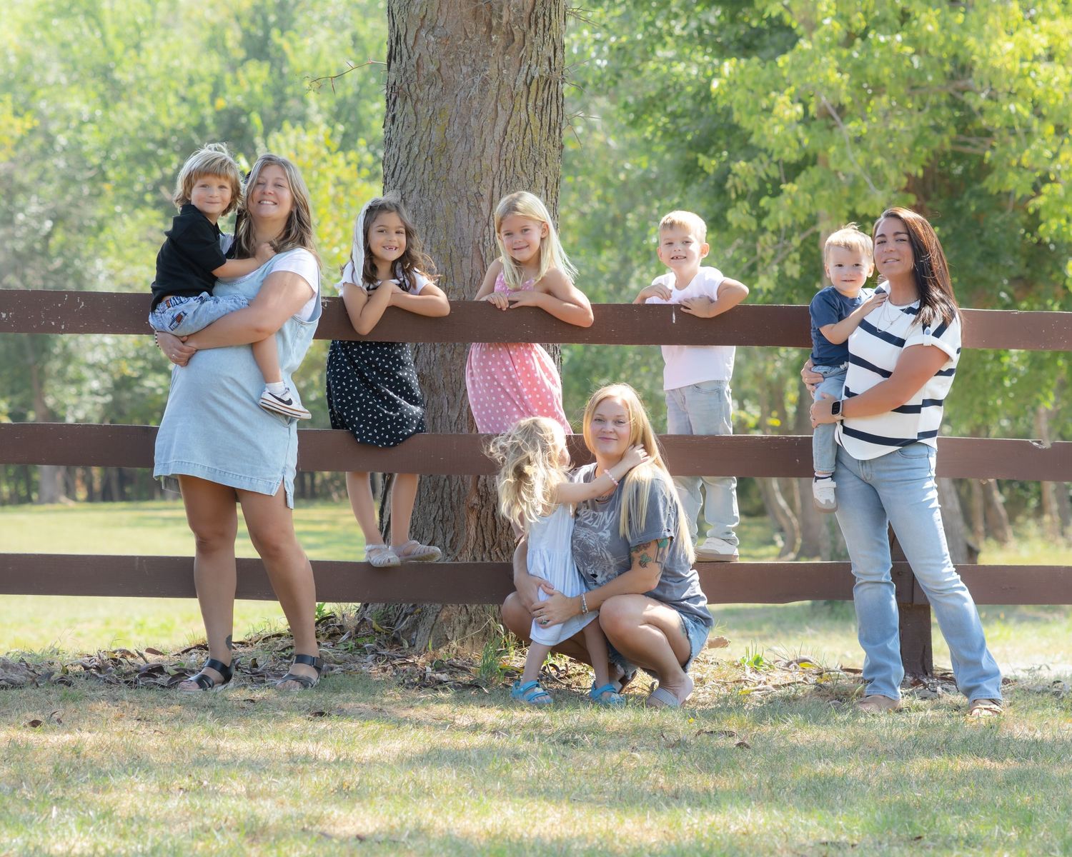 Family group photo with children and adults posing by a wooden fence surrounded by lush green trees outdoors.