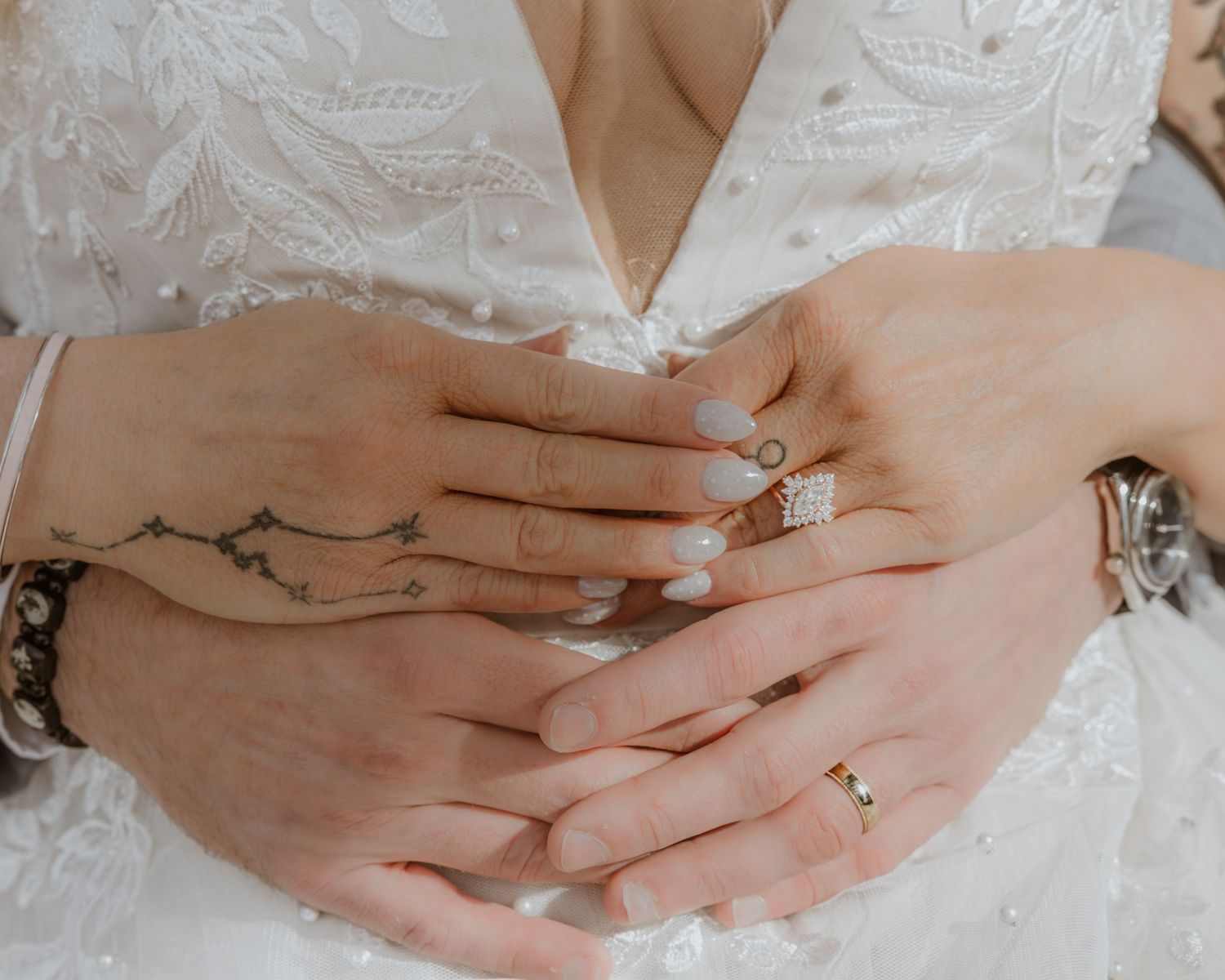 Bride and groom hands clasped together showing wedding rings, tattoo, and lace wedding dress.