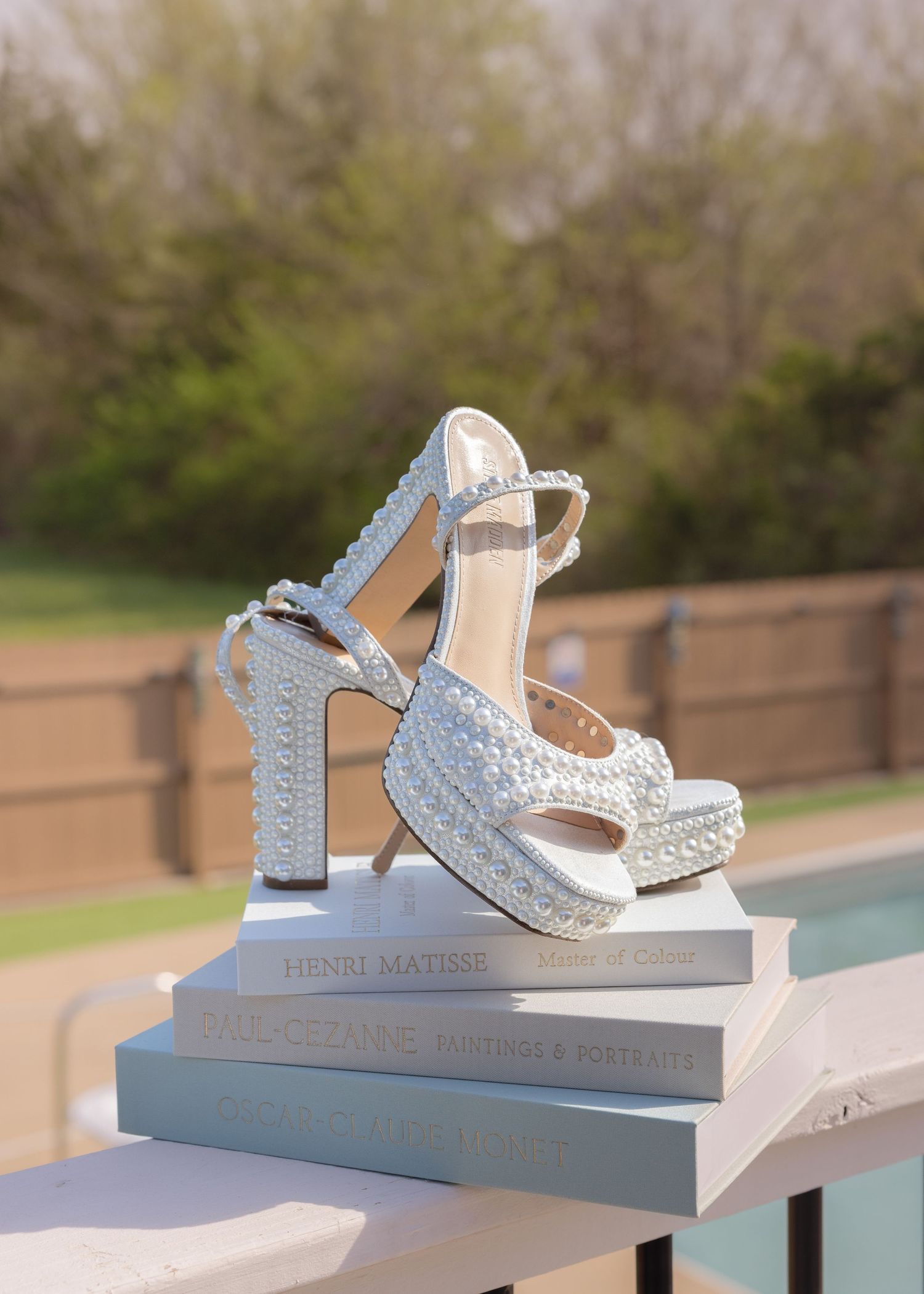 Pearl-embellished white bridal platform heels displayed on stacked books near a pool.