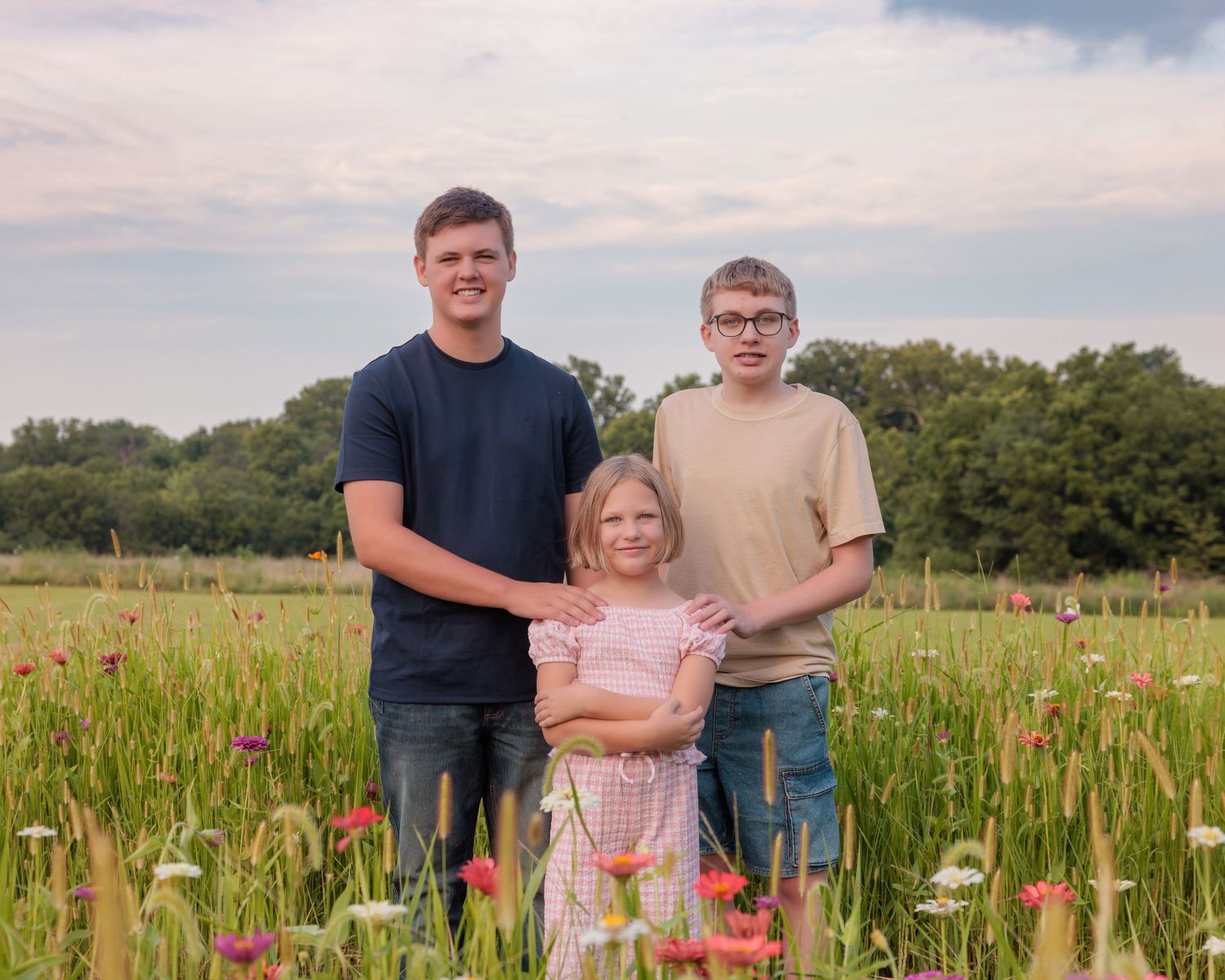 Three siblings posing in a vibrant wildflower meadow with lush green trees and a cloudy sky backdrop.