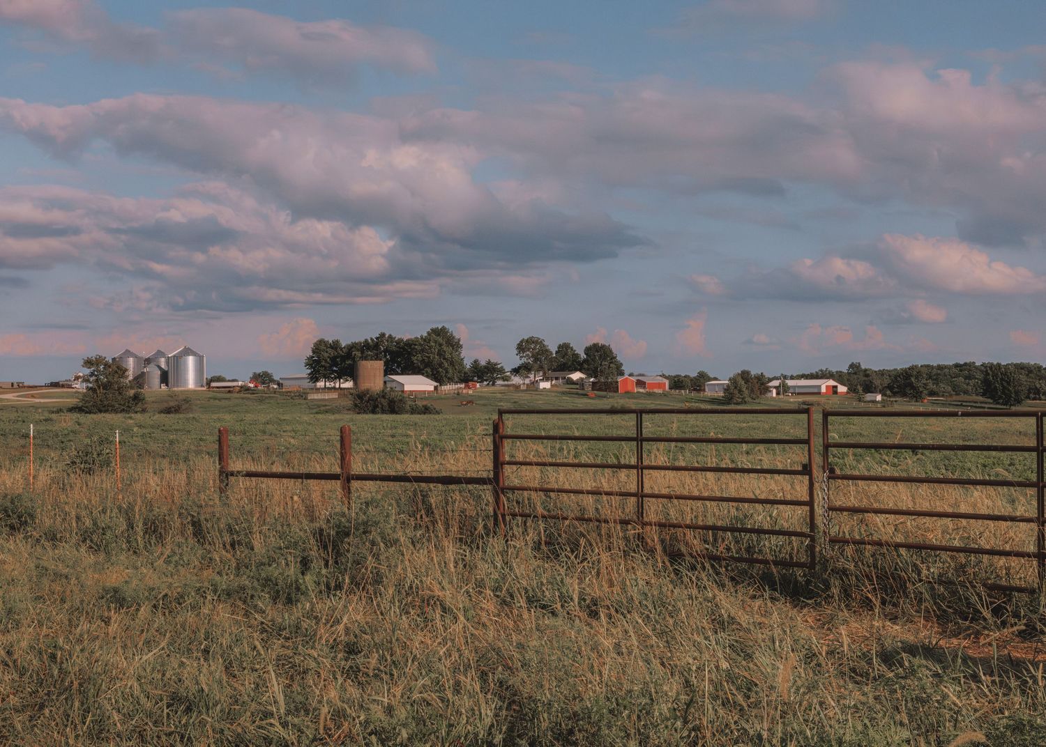 A rustic metal farm gate overlooks a vast green pasture with red barns, grain silos, and trees under a cloudy sky.
