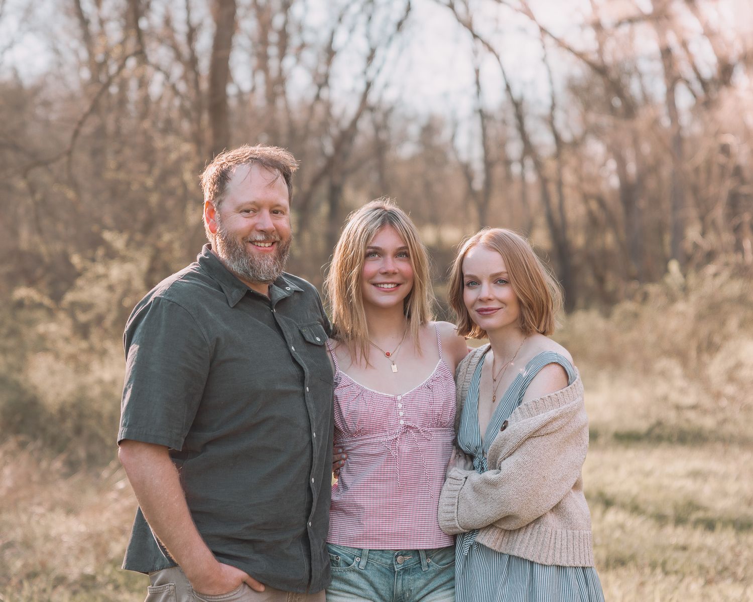 A man and two girls smile together outdoors in a golden-hour nature setting with trees in the background.