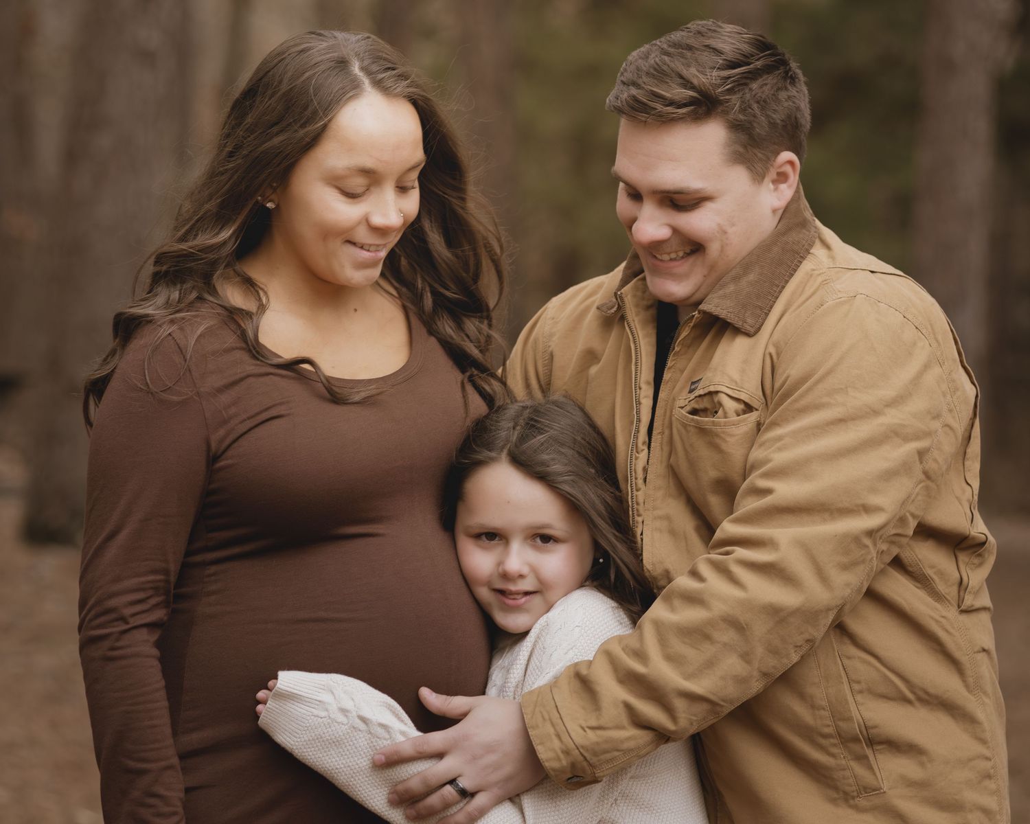 Pregnant woman in brown top with husband and daughter posing for outdoor maternity photo shoot in forest.