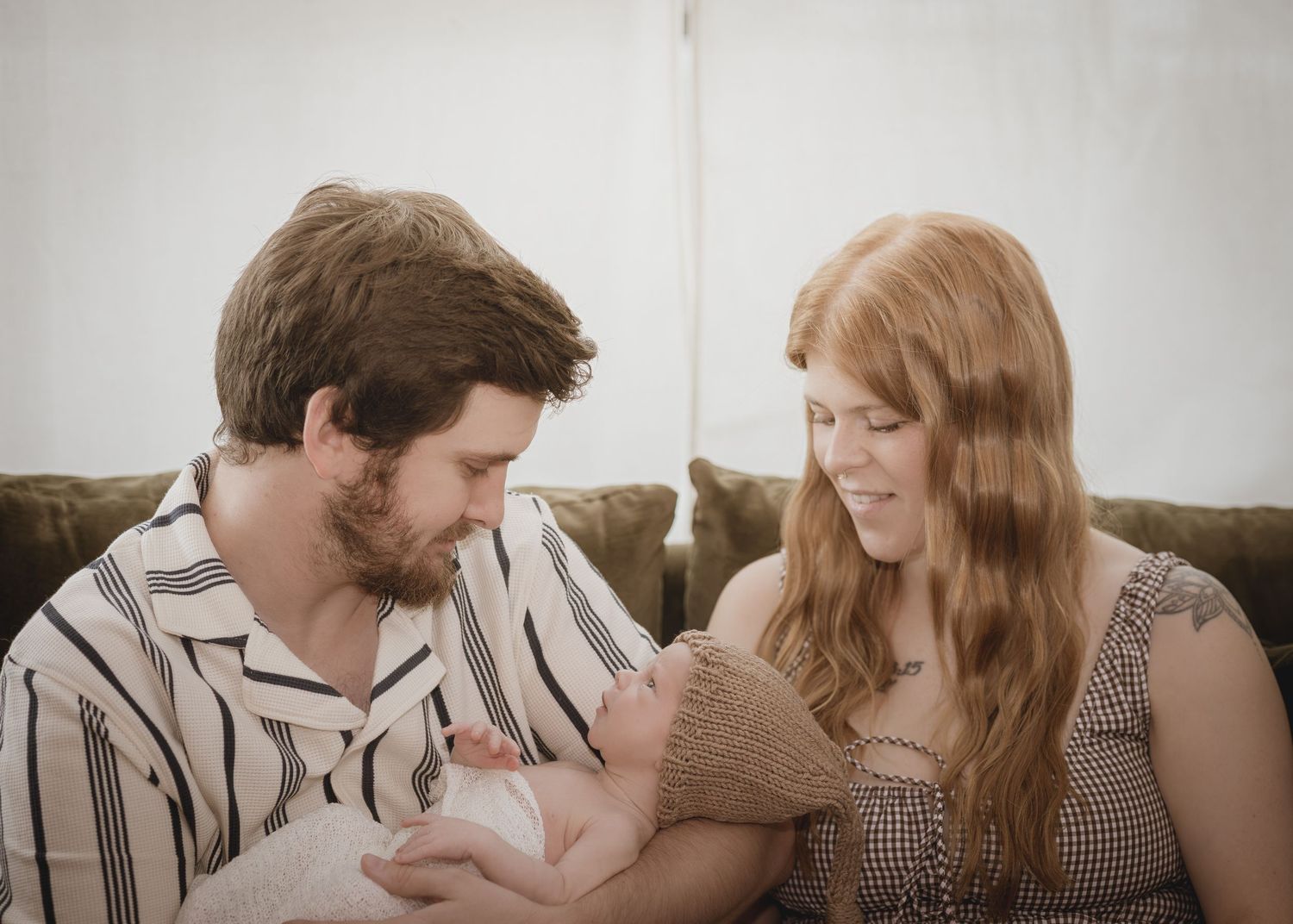 A couple smiles lovingly at their newborn baby while sitting on a couch.
