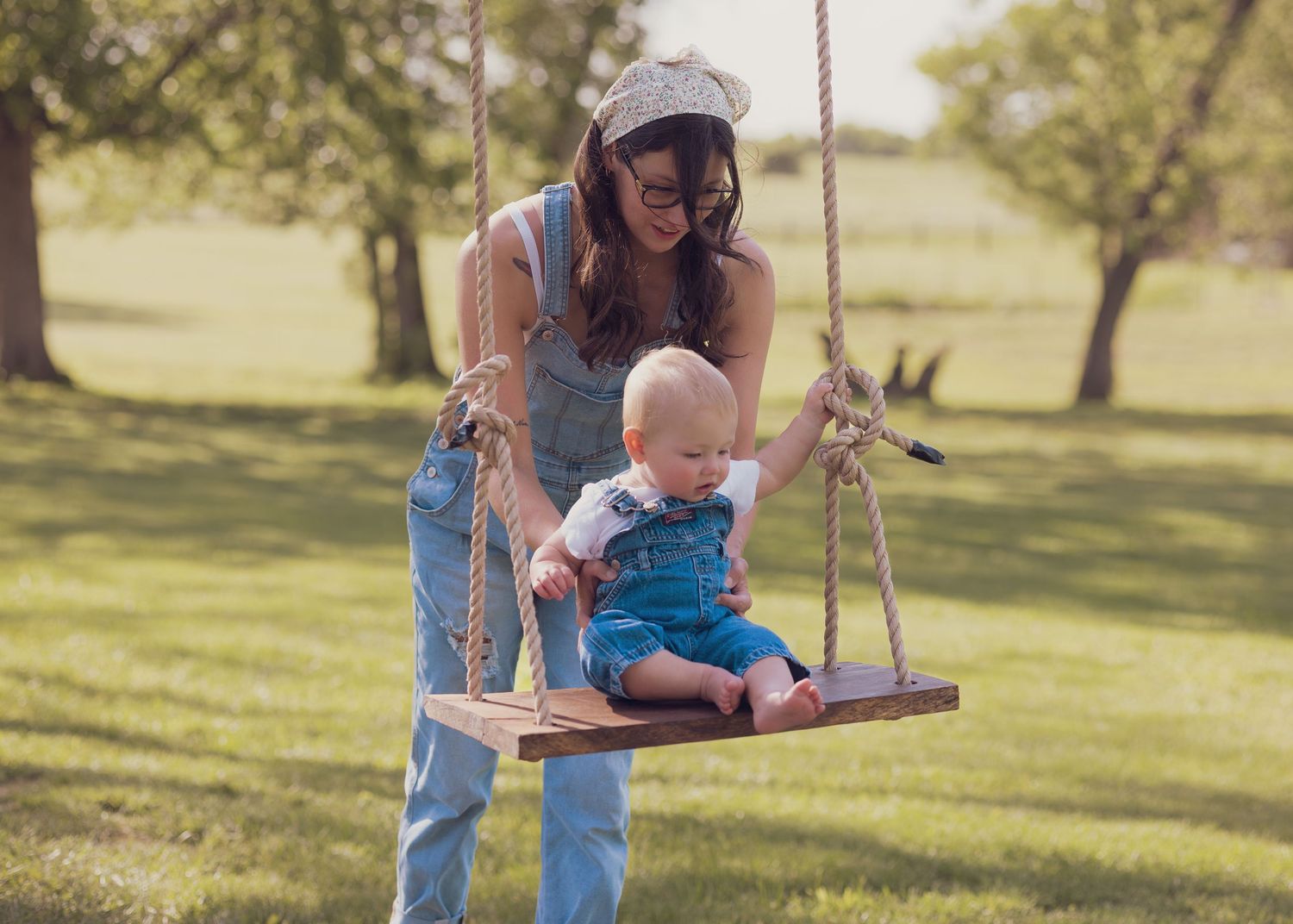 Mother helping baby sit on wooden rope swing outdoors in green park setting.