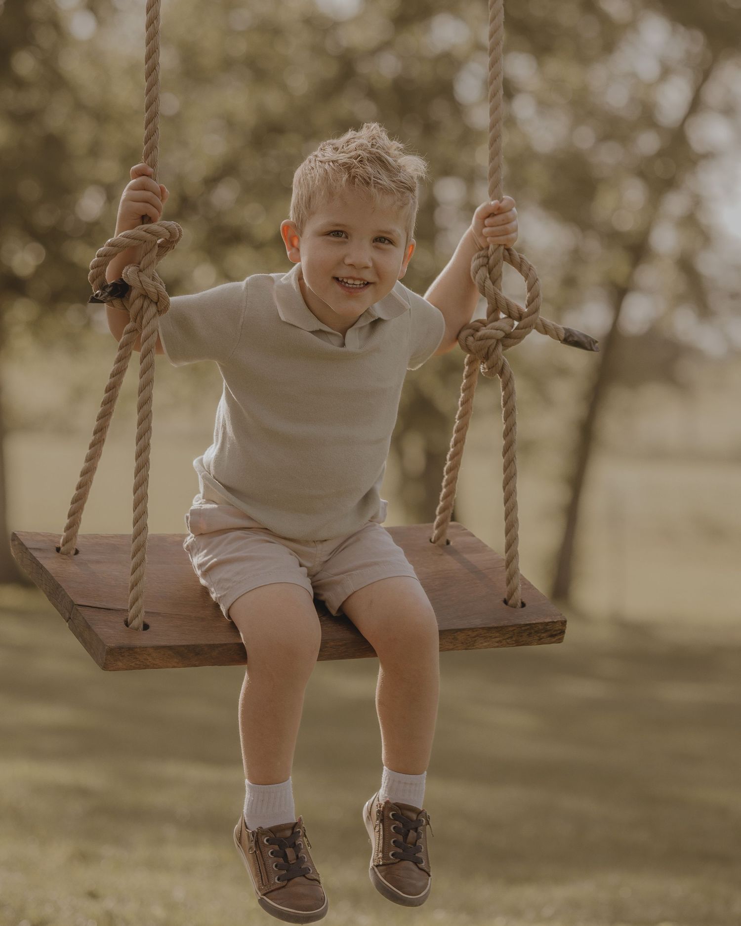 A happy young boy in a polo shirt sits smiling on a wooden rope swing in a sunny park.