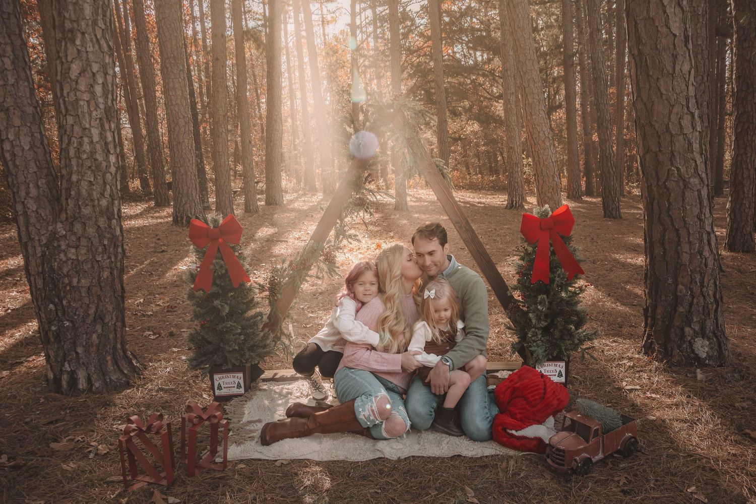 A family sits on a blanket in a sunlit pine forest decorated with red Christmas decor and presents.