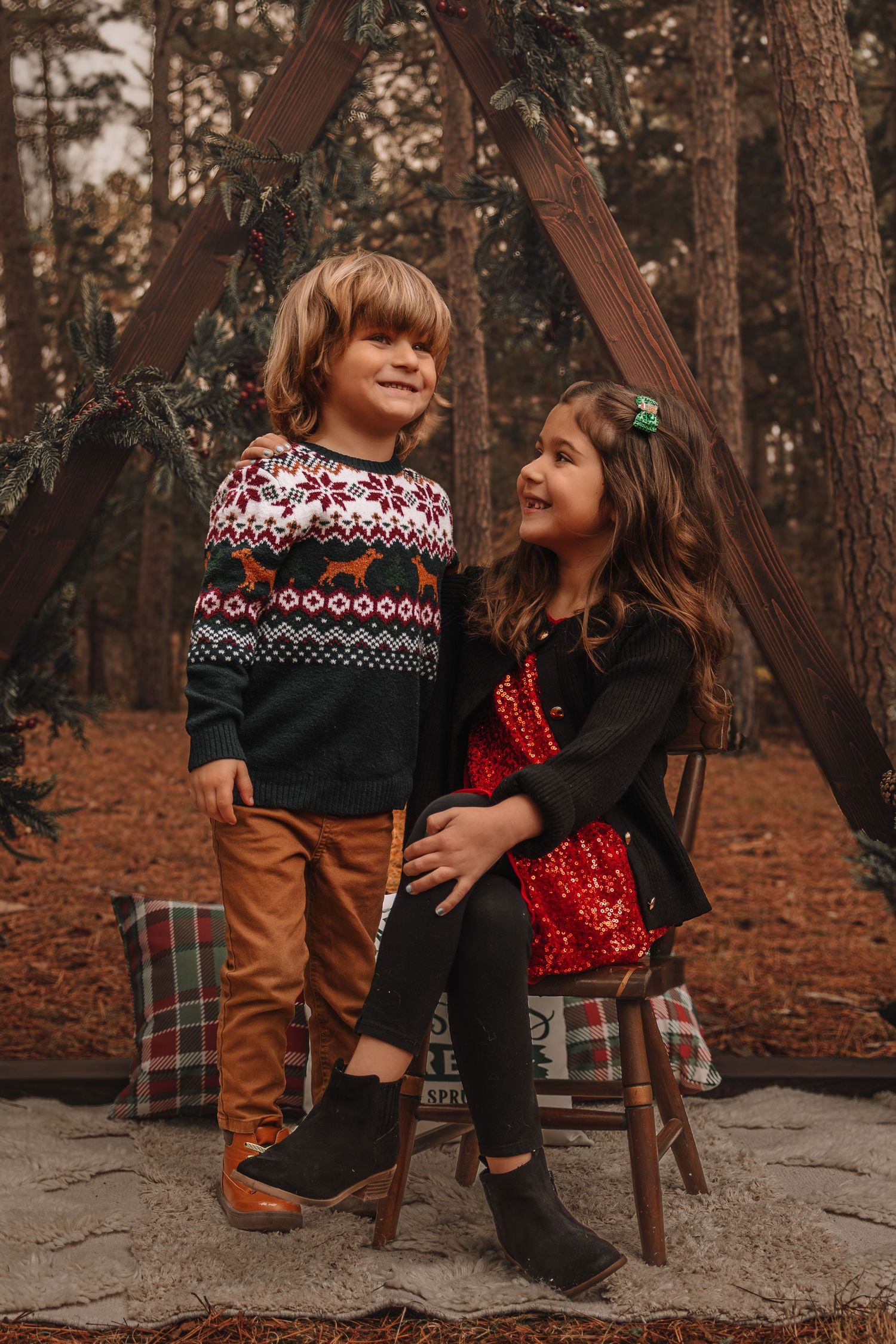 Two children in winter sweaters sit together on a rock in a pine forest during golden hour.