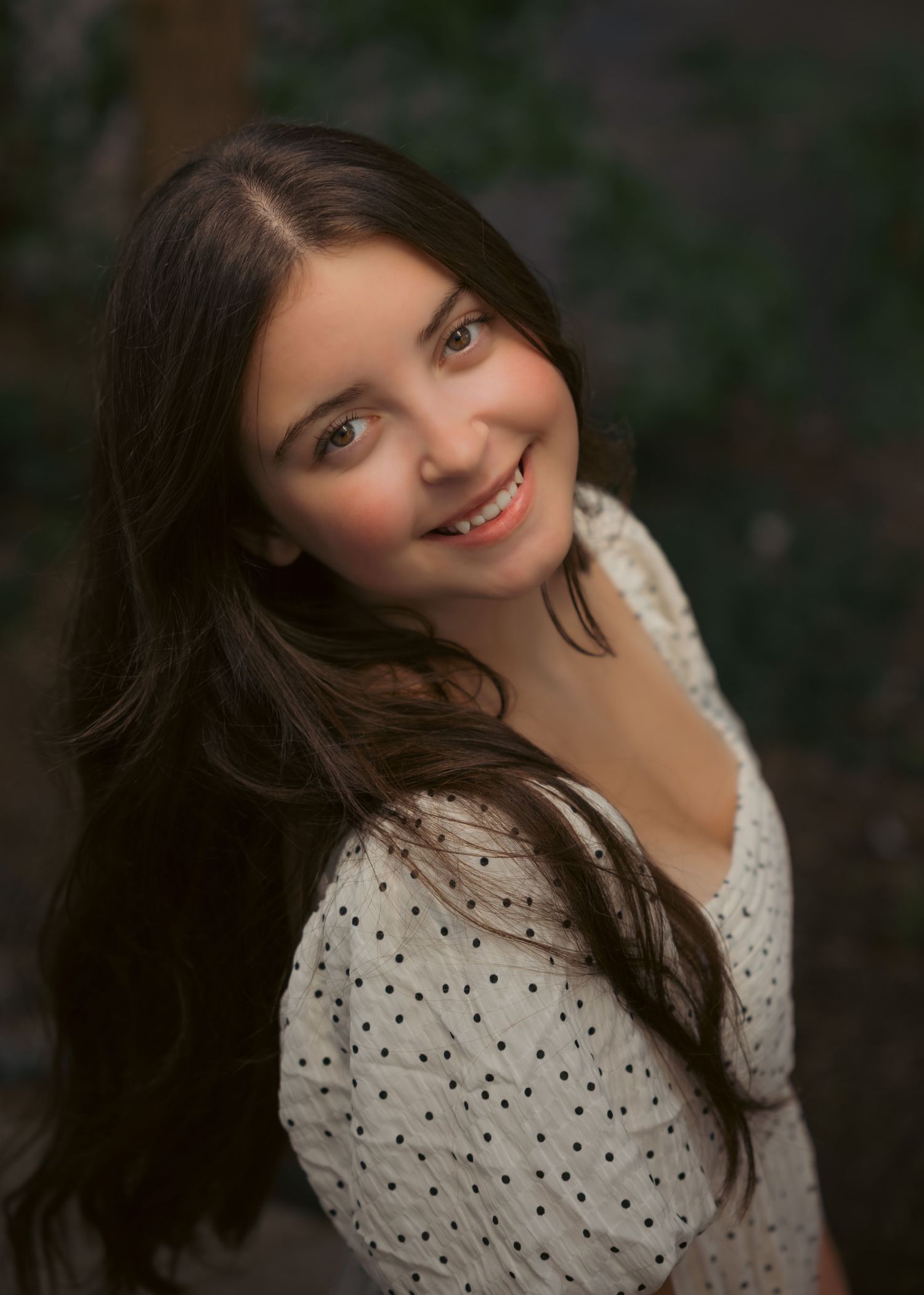 Young woman with long dark hair smiling outdoors, wearing a white polka dot top in a portrait photo.