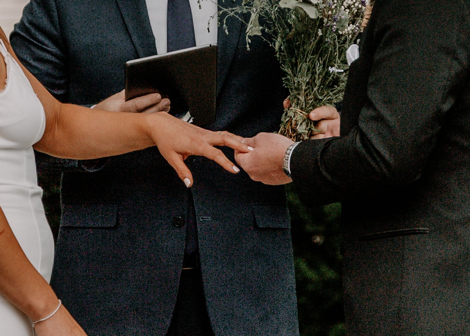 Close up of hands during ring exchange ceremony with bouquet in background.