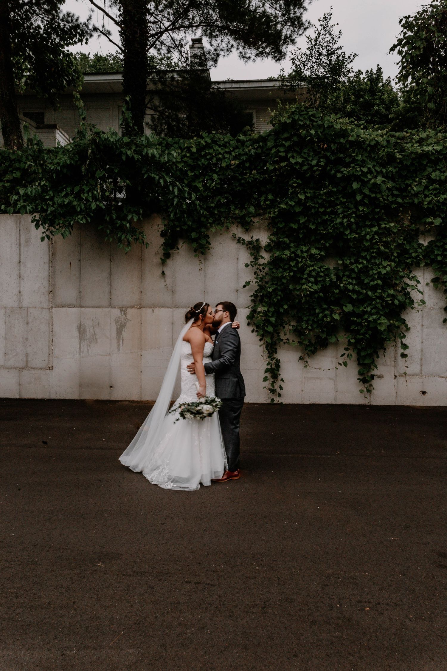 Romantic wedding couple poses against an ivy-covered wall in elegant attire.