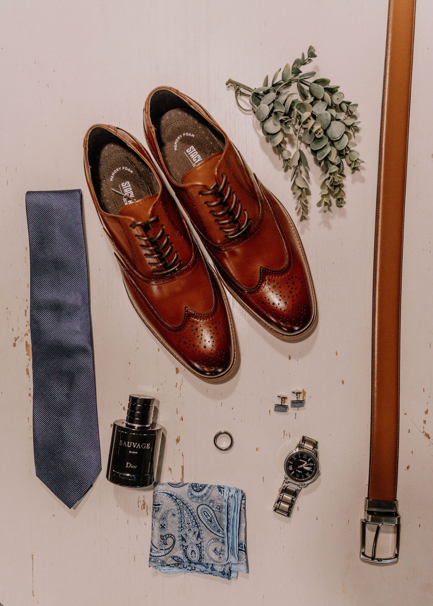 Wedding accessories including brown leather shoes, blue tie, watch and cologne arranged on white surface.