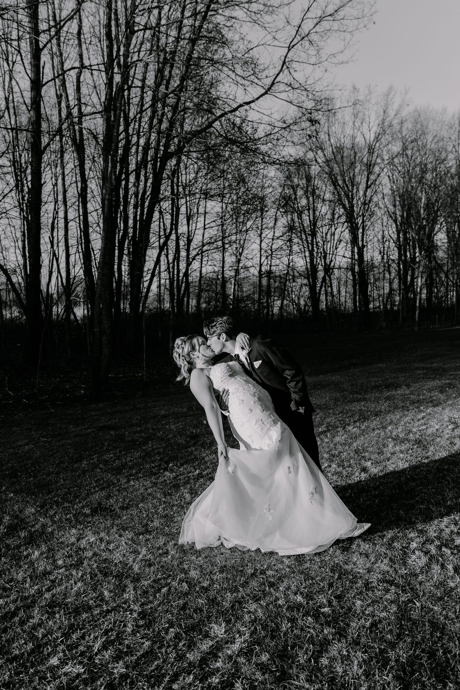 A dramatic black and white photo of newlyweds embracing against a backdrop of bare trees.