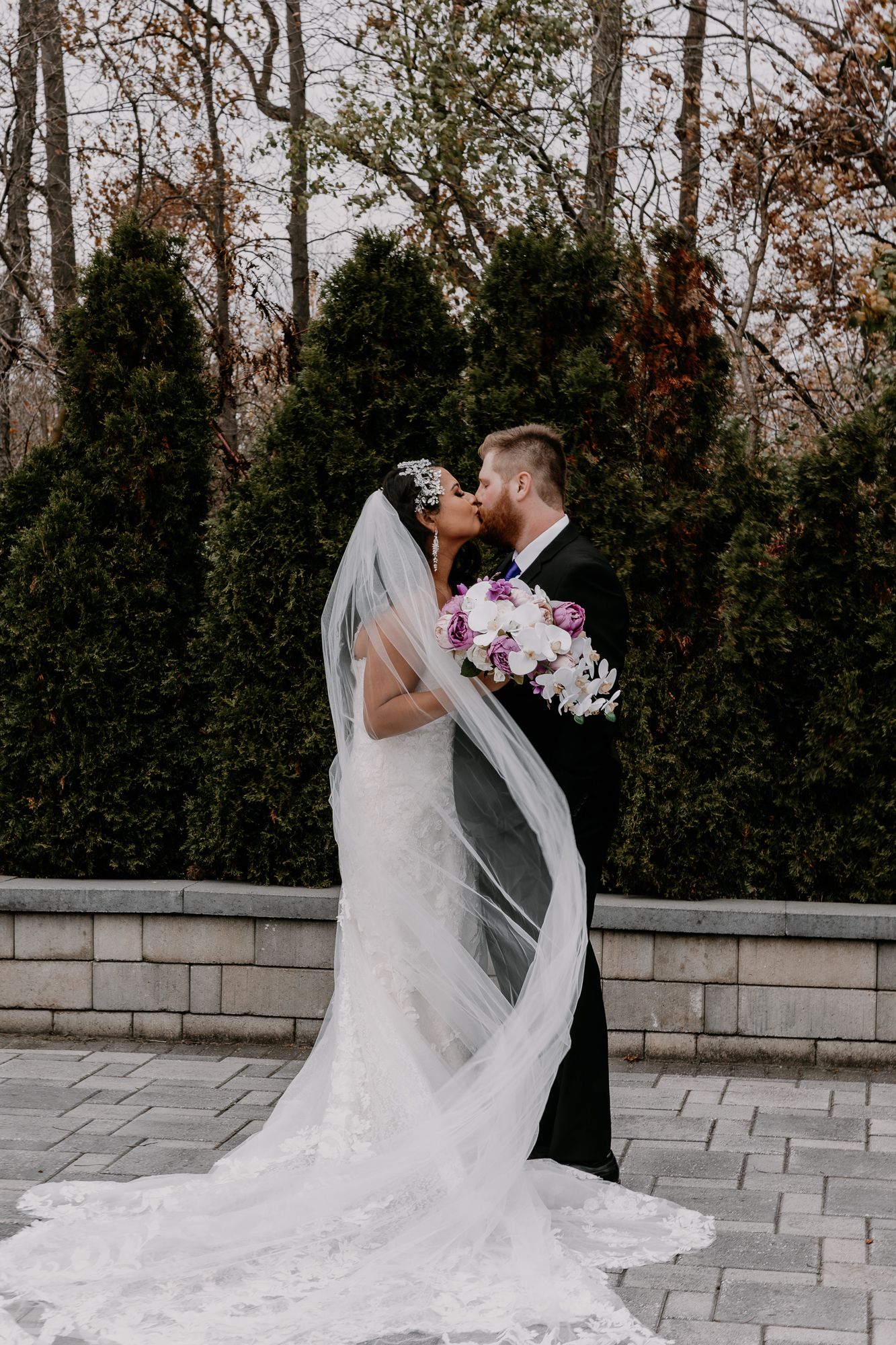 Newlyweds share an intimate moment in a garden with the bride's veil flowing in the wind.