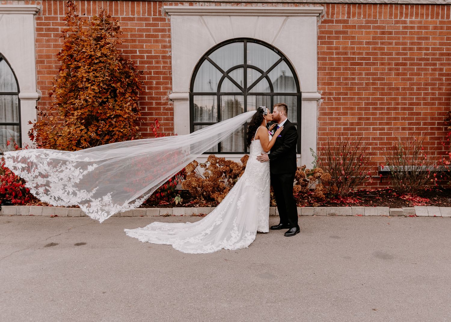 Long lace veil flows in the wind as newlyweds pose by brick building with arched window.