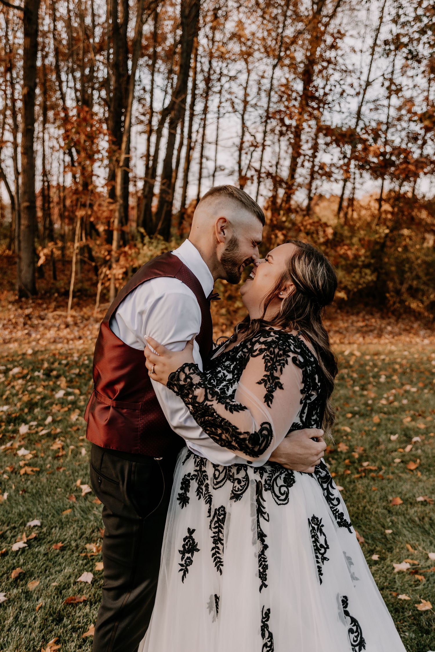 Intimate moment between newlyweds in autumn forest wearing burgundy vest and black lace wedding dress.