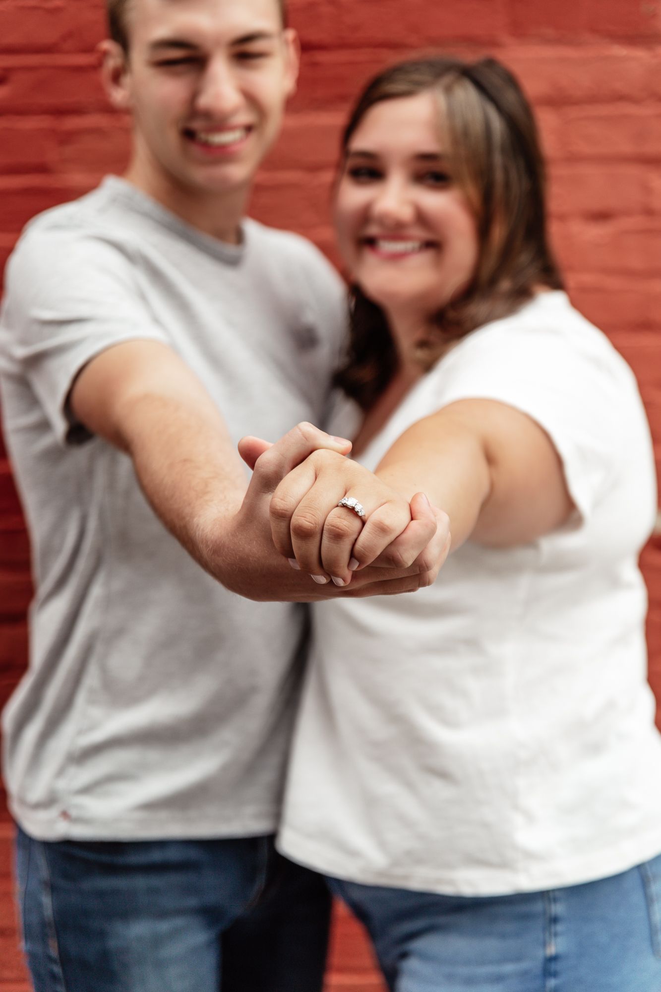 Couple shows off engagement ring with hands clasped together against red brick wall.