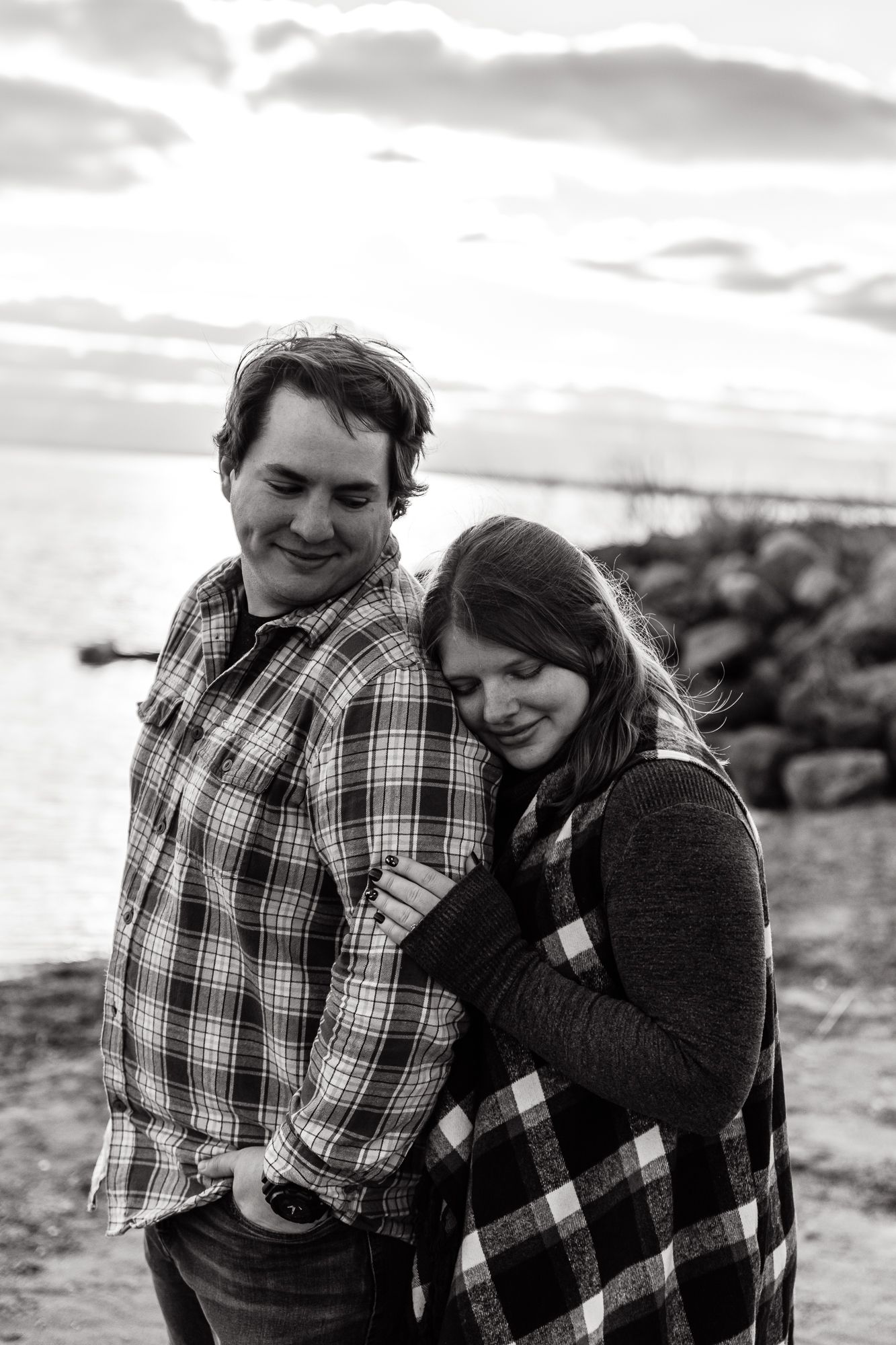 A couple in plaid clothing embraces on a rocky beach during sunset.