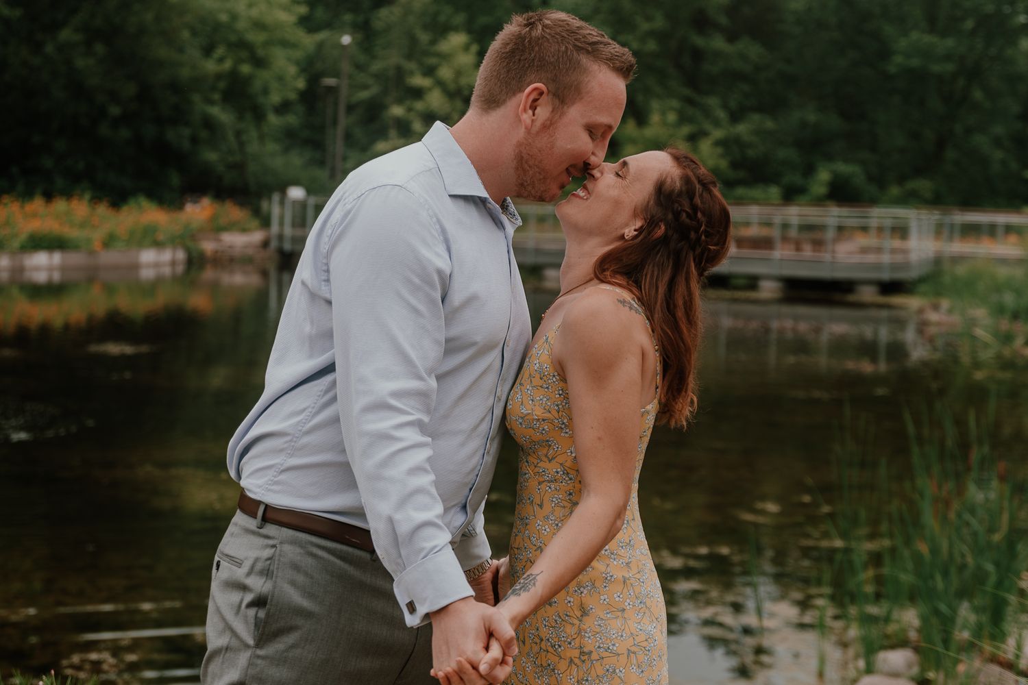 A couple shares a romantic kiss by a pond surrounded by lush summer gardens.