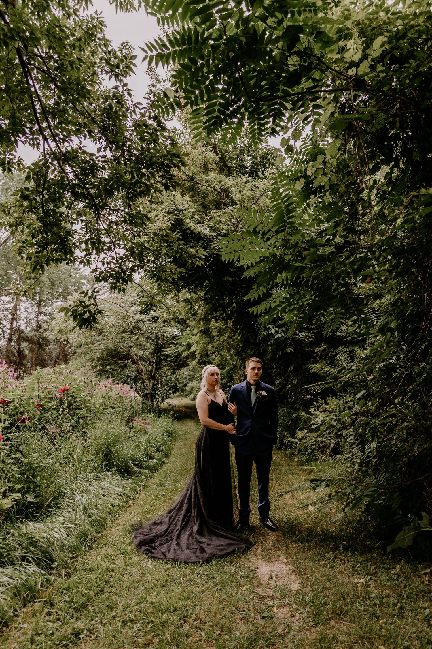 Couple in black wedding attire posing on a lush garden path surrounded by green trees and wildflowers.