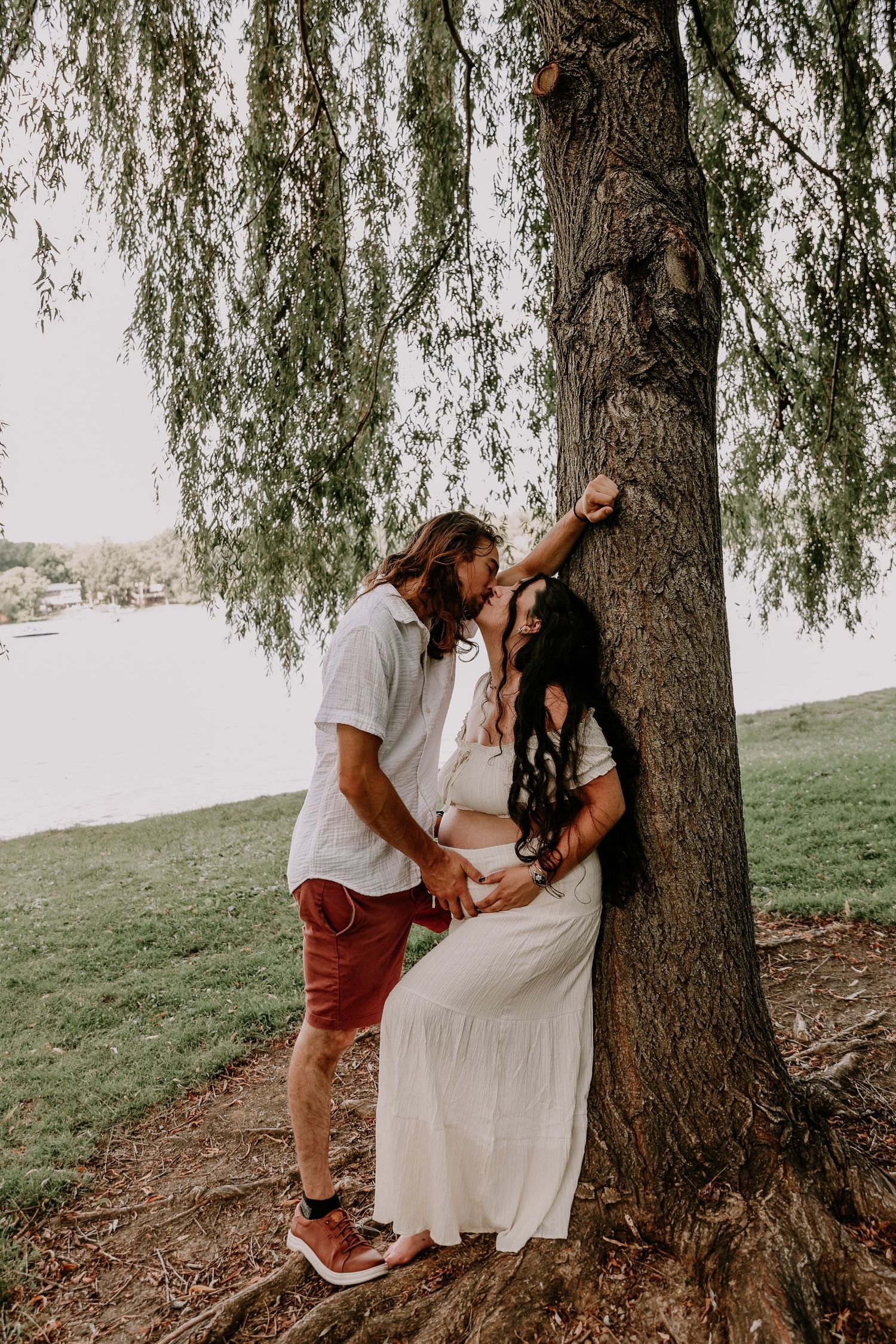 A couple shares a tender kiss under a weeping willow tree, the woman in a white dress cradling her baby bump.