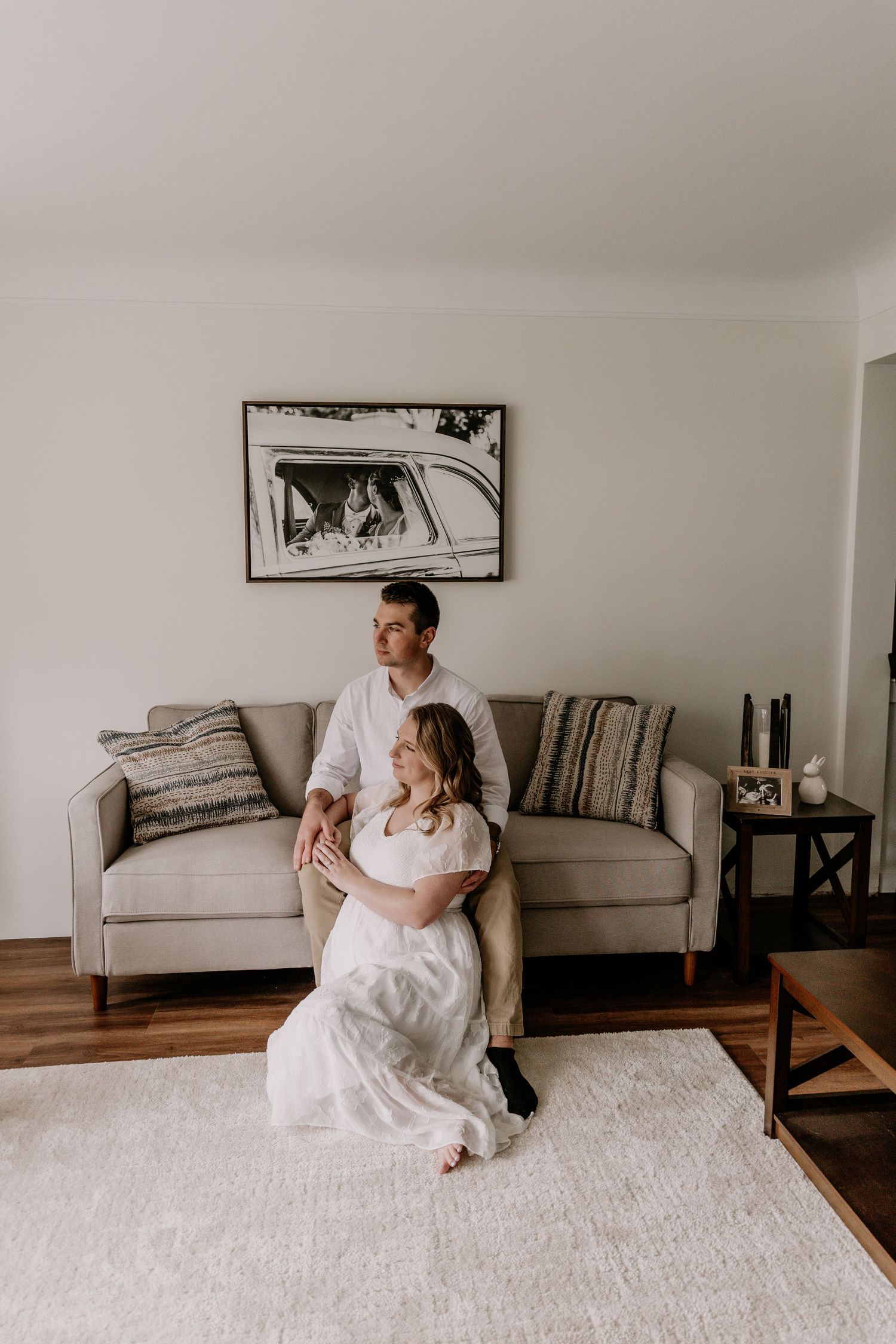 Couple sitting together on living room floor in front of gray sofa, woman in white dress, man in white shirt, cozy home.