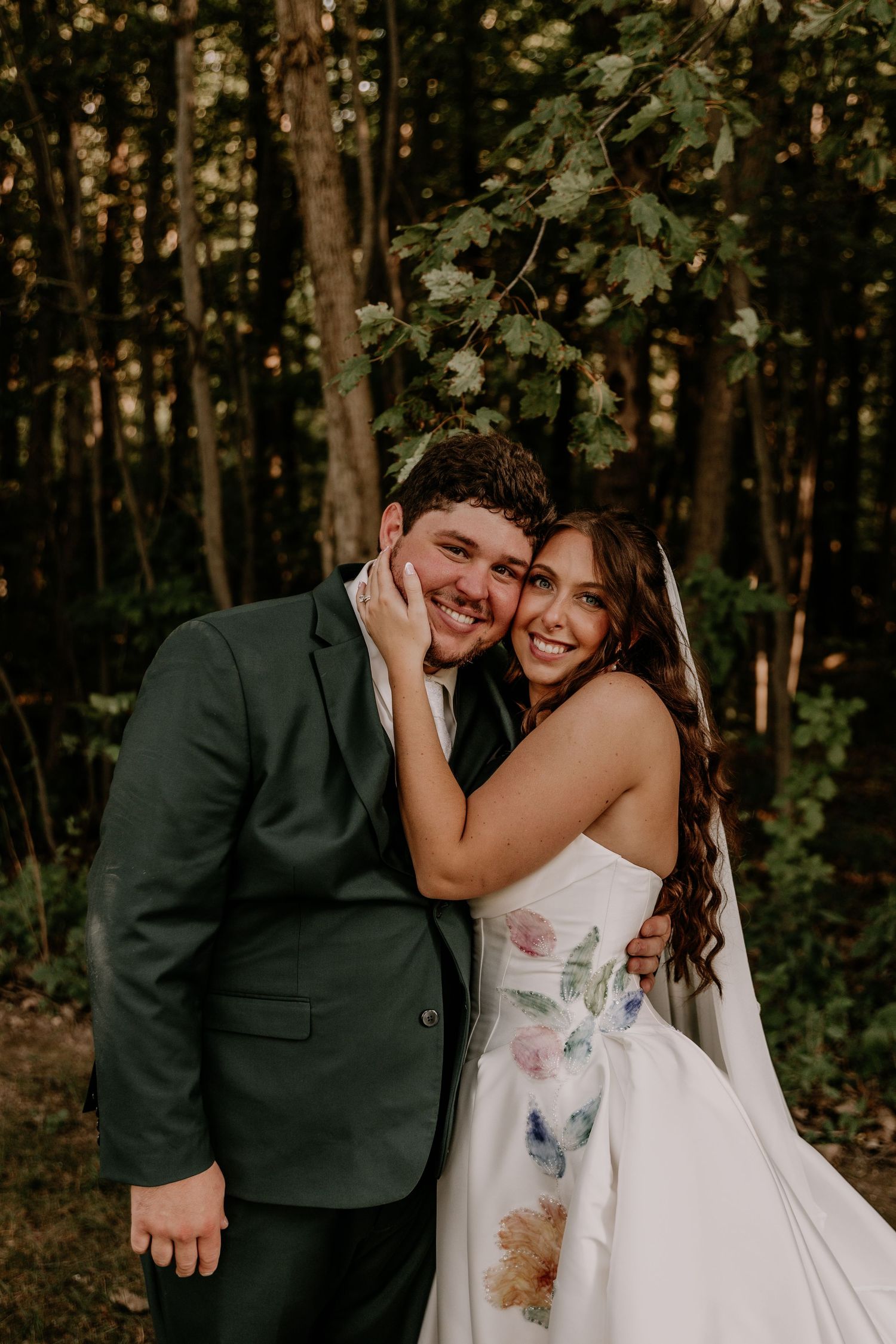 A smiling bride in a floral gown embraces a groom in a dark green suit outdoors in a wooded setting.
