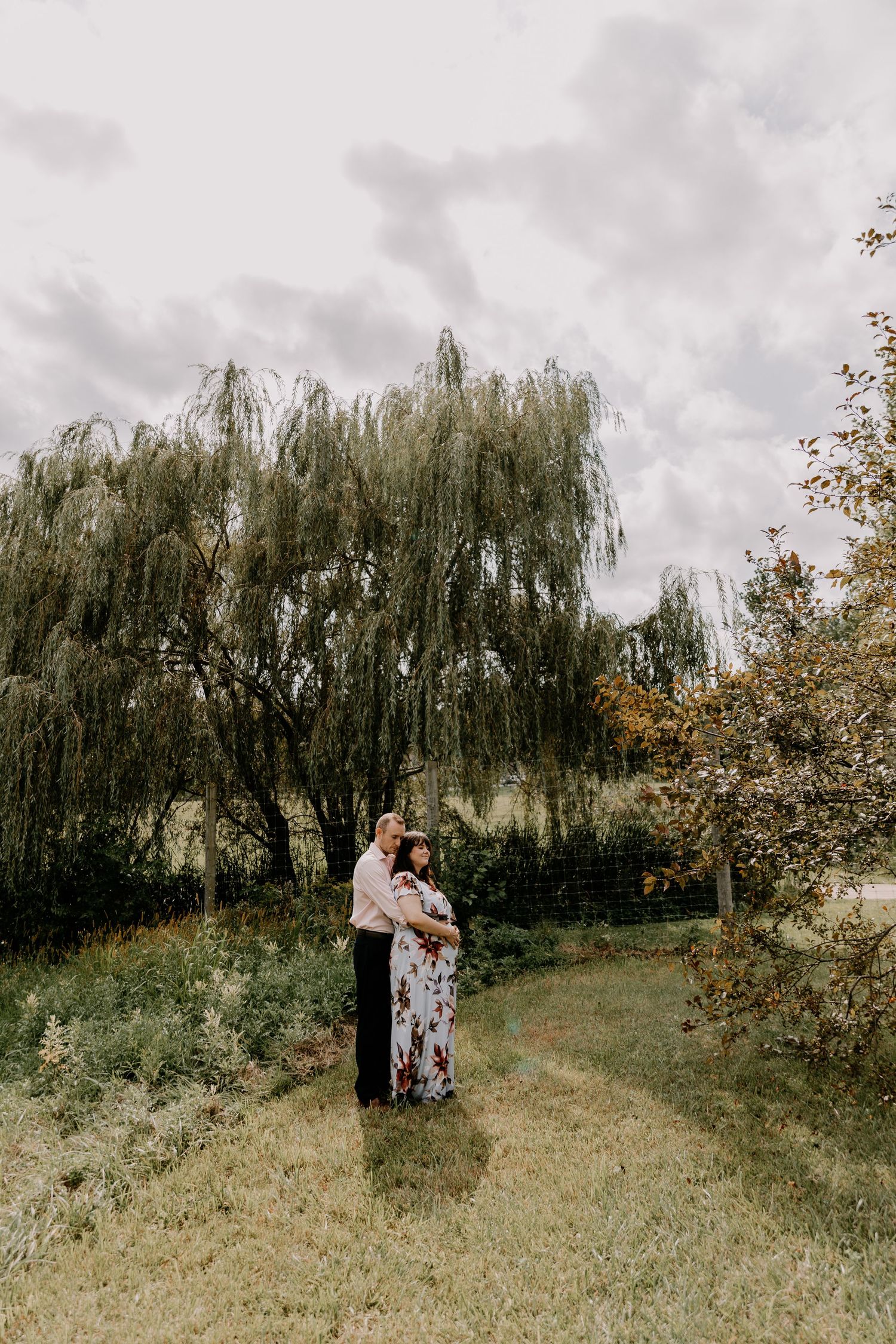A romantic couple embraces in a picturesque orchard with weeping willow trees and autumn foliage in the background.