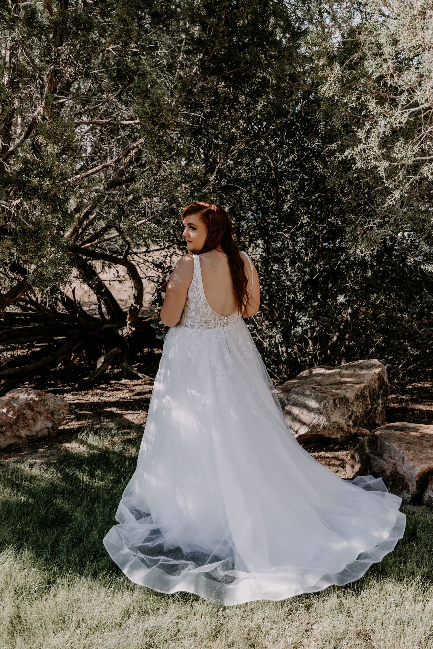 A flowing white wedding dress trails elegantly across green grass under shaded trees.