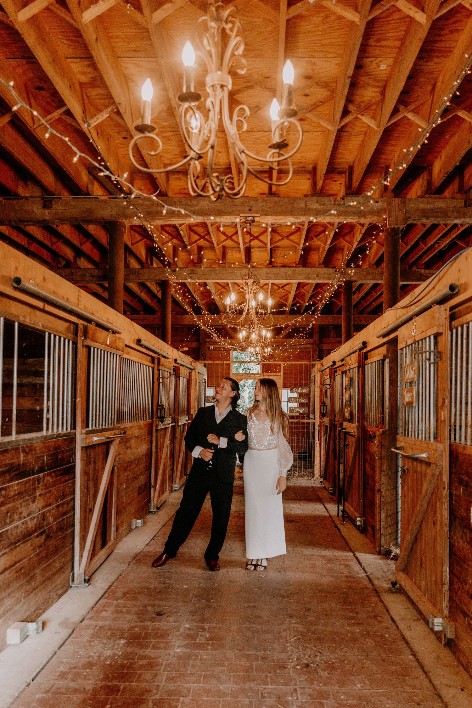 A rustic barn venue interior with elegant chandeliers and wooden stalls decorated for a wedding ceremony.