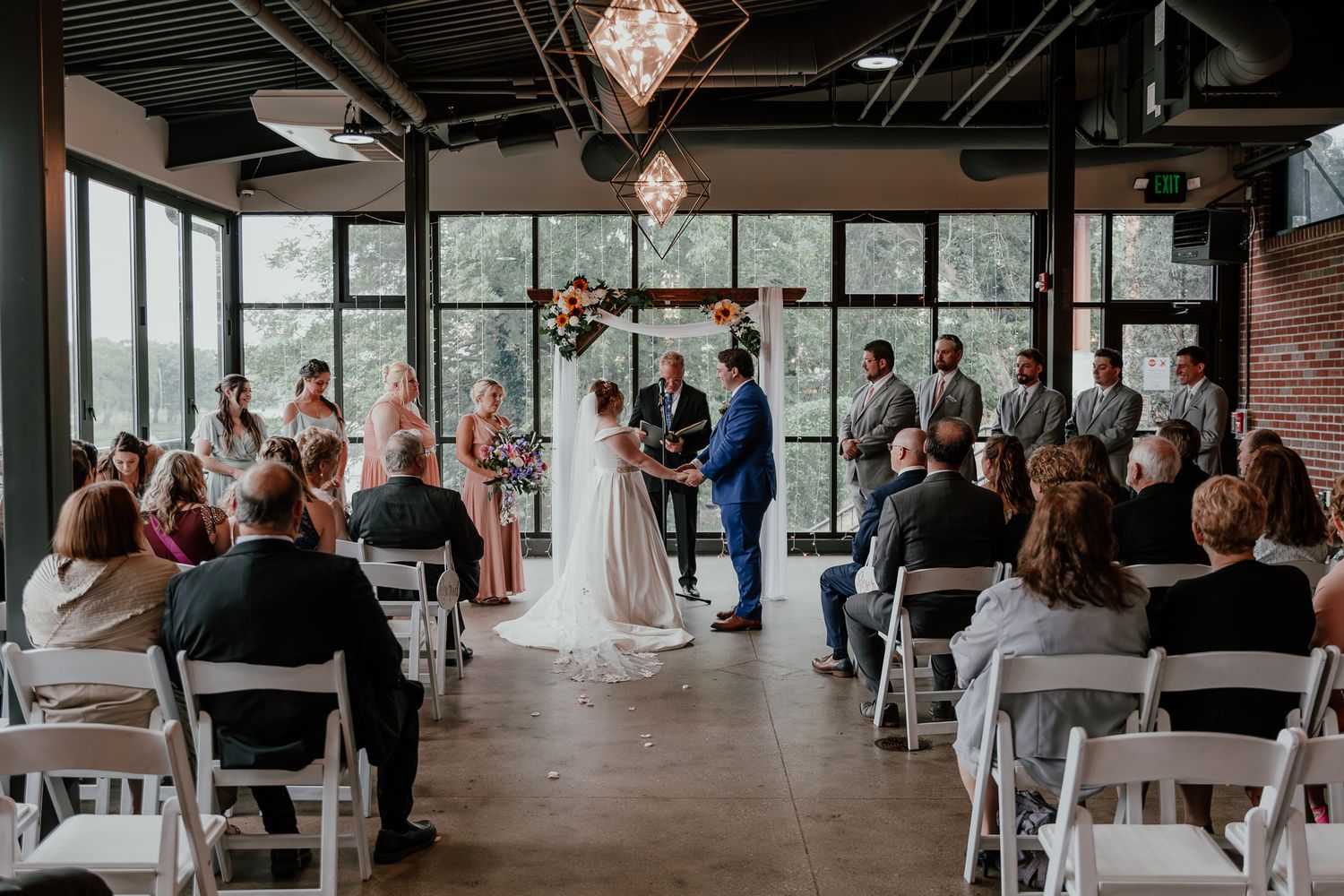 Modern indoor wedding ceremony with guests seated in white chairs watching couple exchange vows.