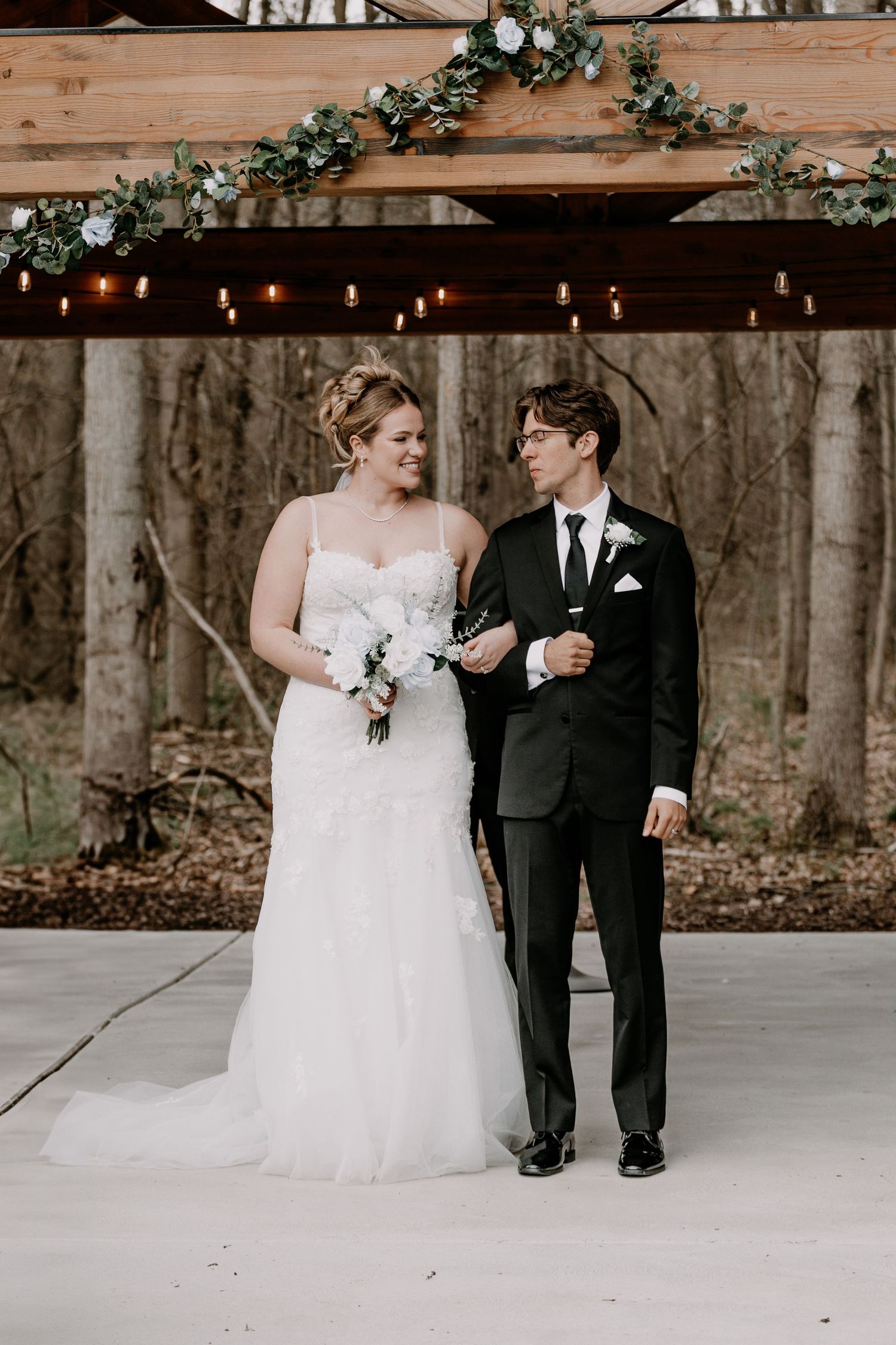 Bride in white gown and groom in black suit pose under wooden arch with greenery.