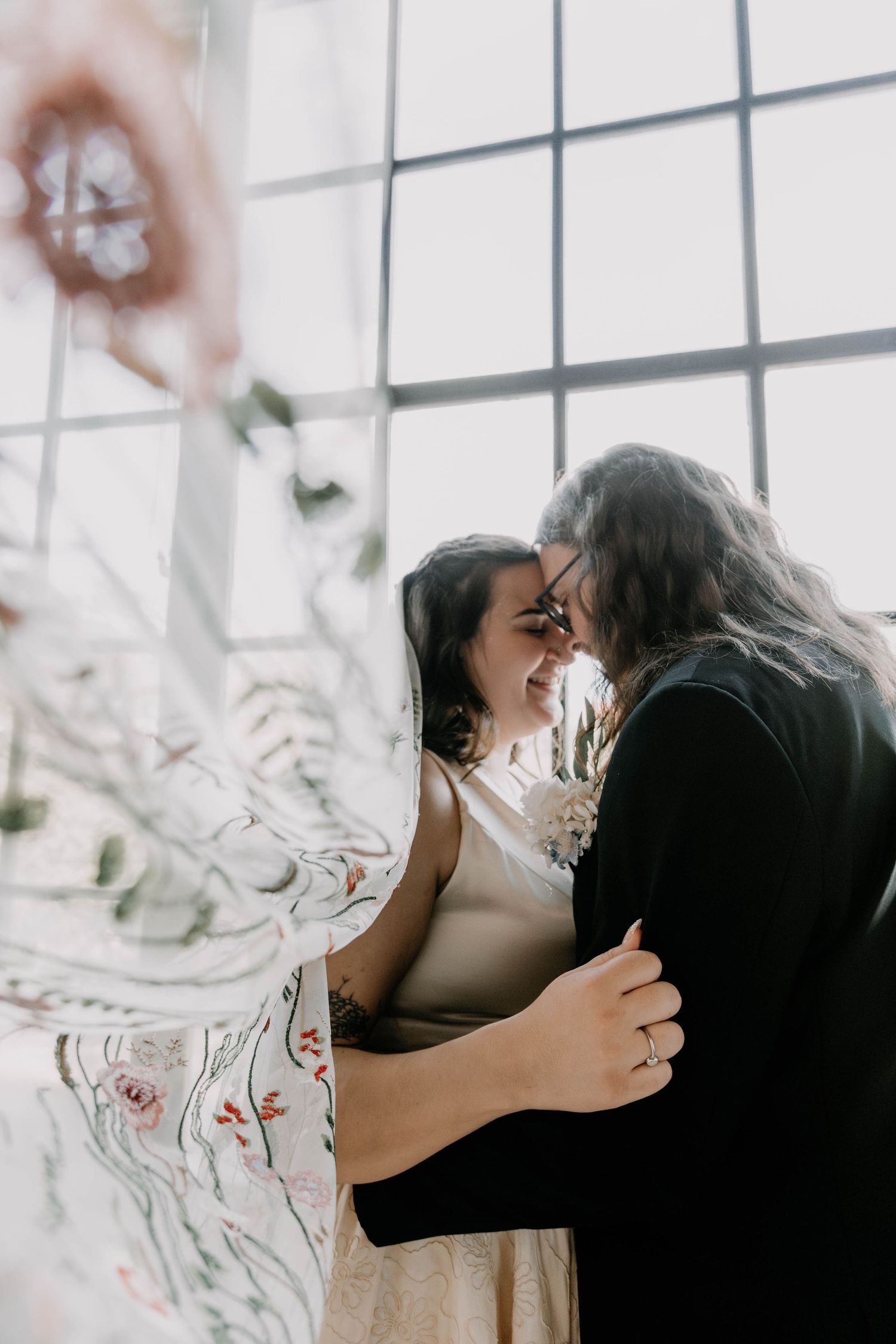 Dreamy candid moment of couple embracing by window with soft light filtering through.