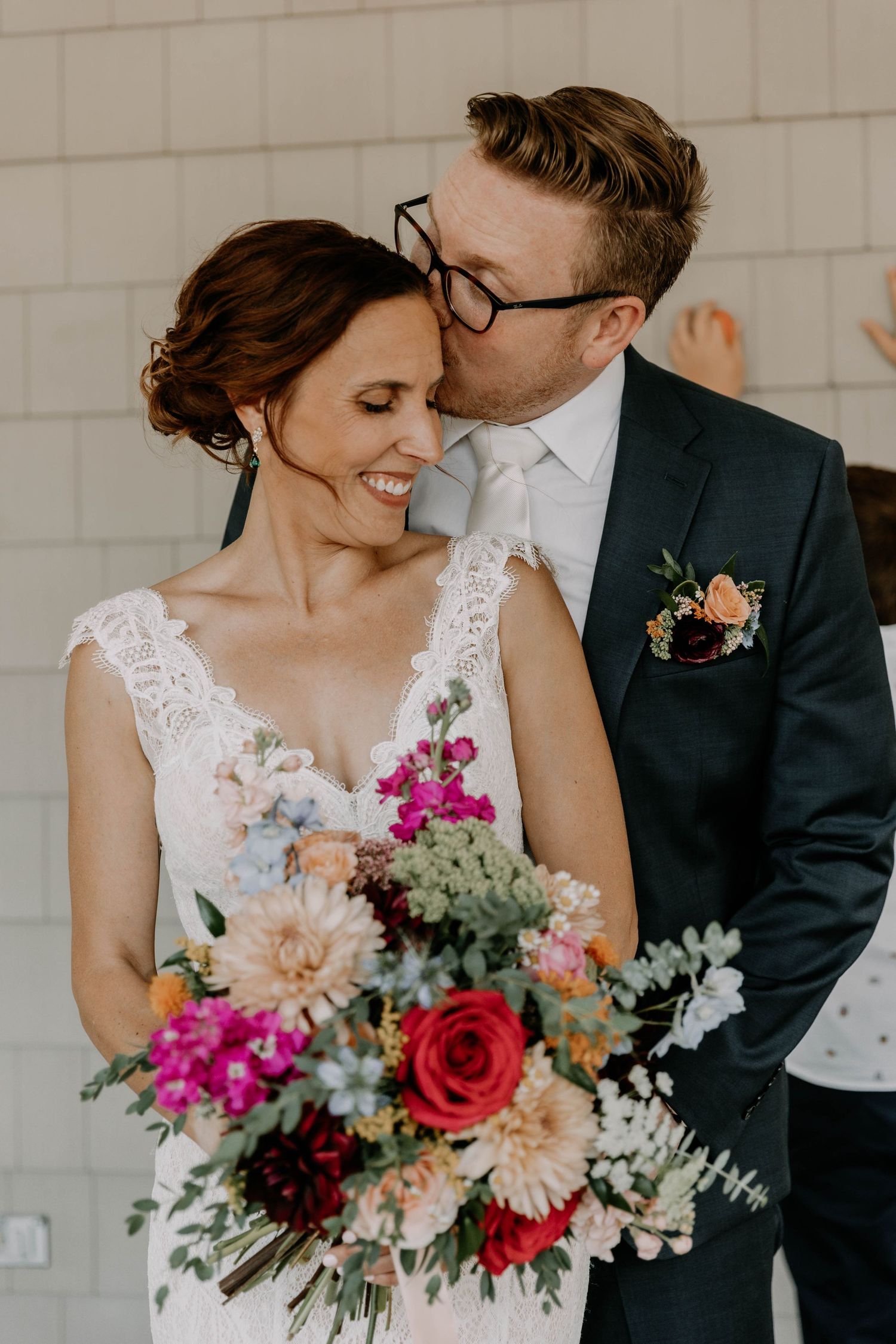 Close-up of wedding couple with vibrant red and pink bouquet against white brick wall.
