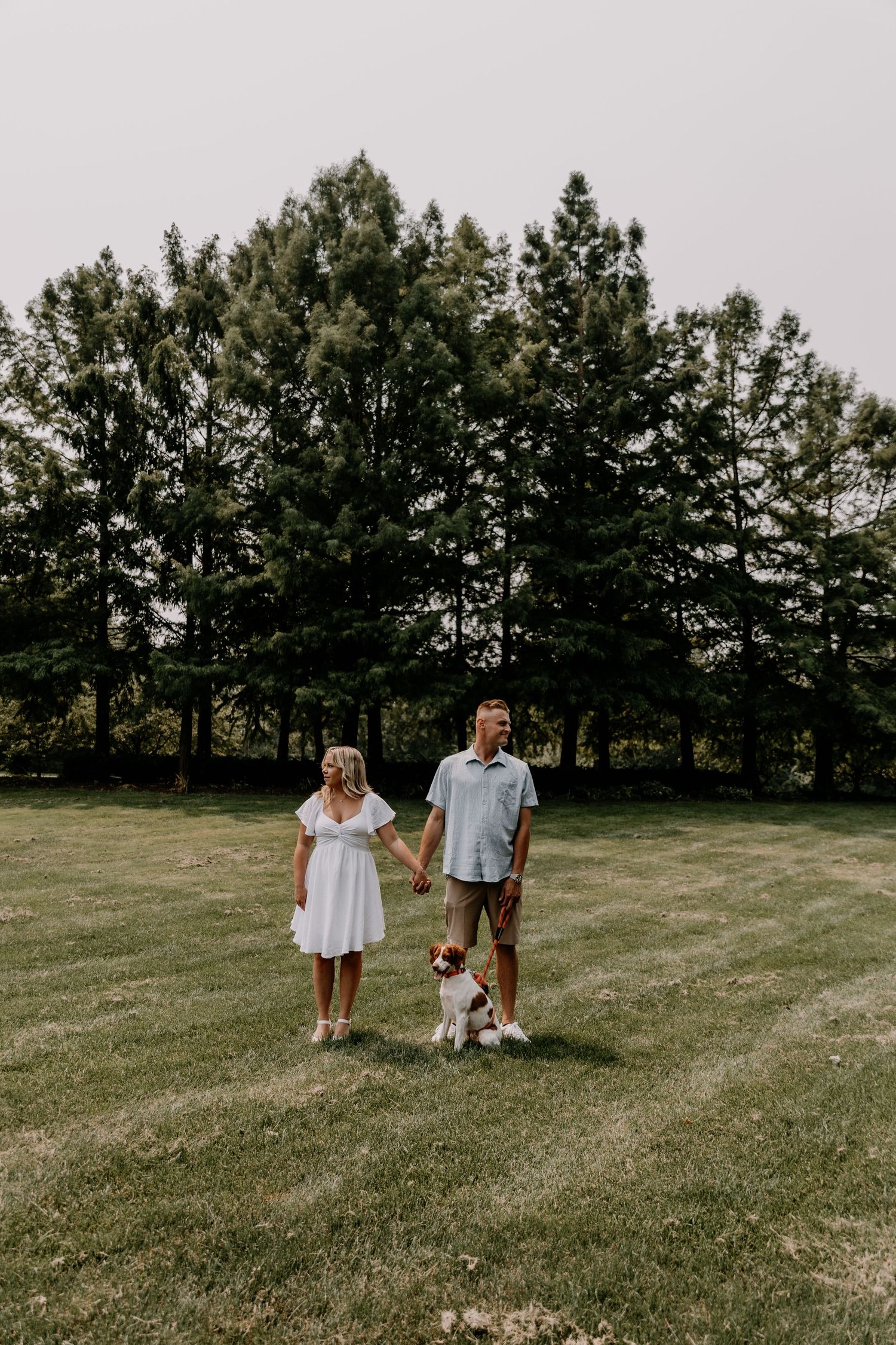 A couple stands on a grassy lawn holding hands with their dog while evergreen trees create a scenic backdrop.