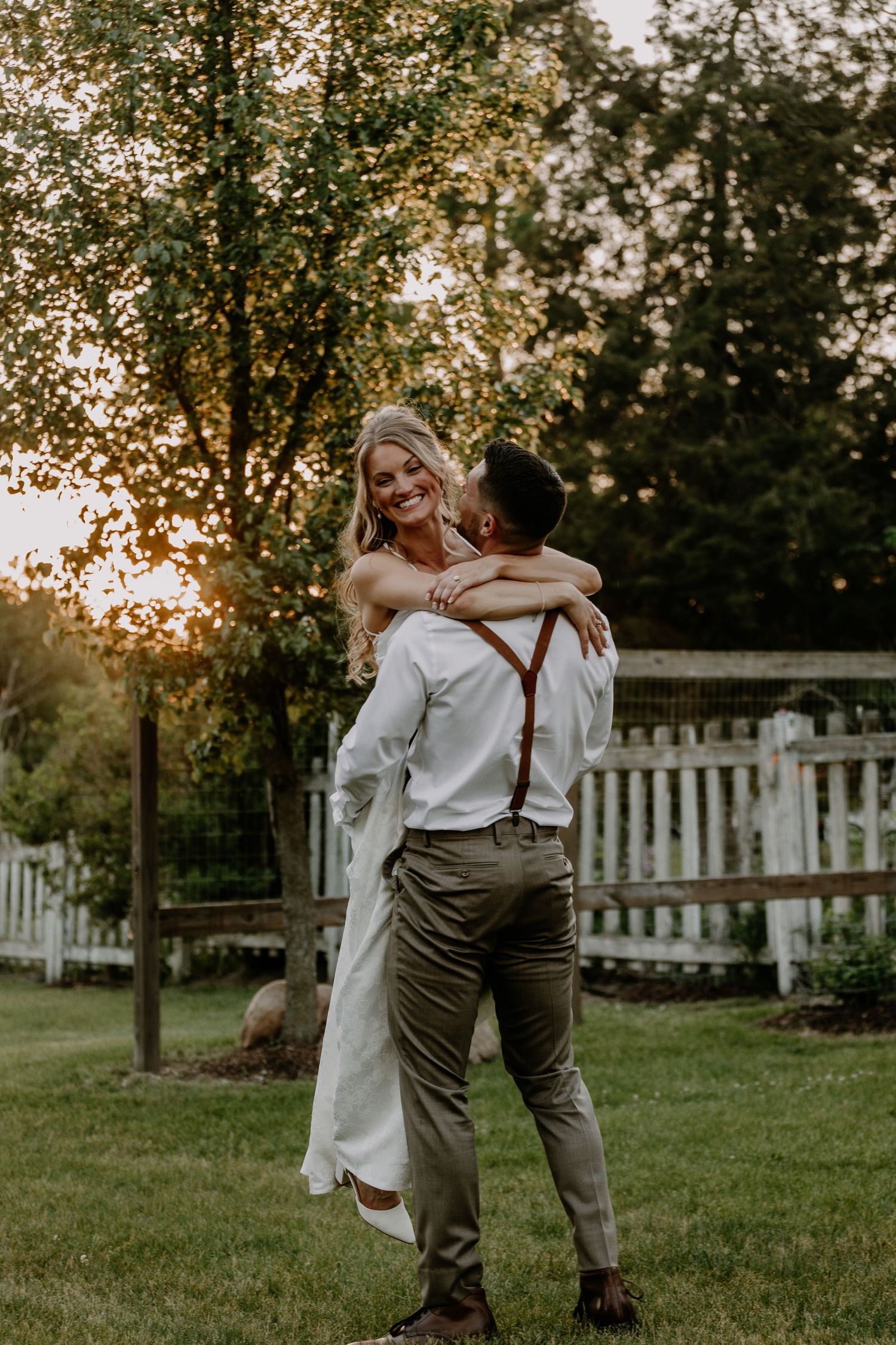 Groom lifting bride in white dress at golden hour in lush garden with white fence backdrop.