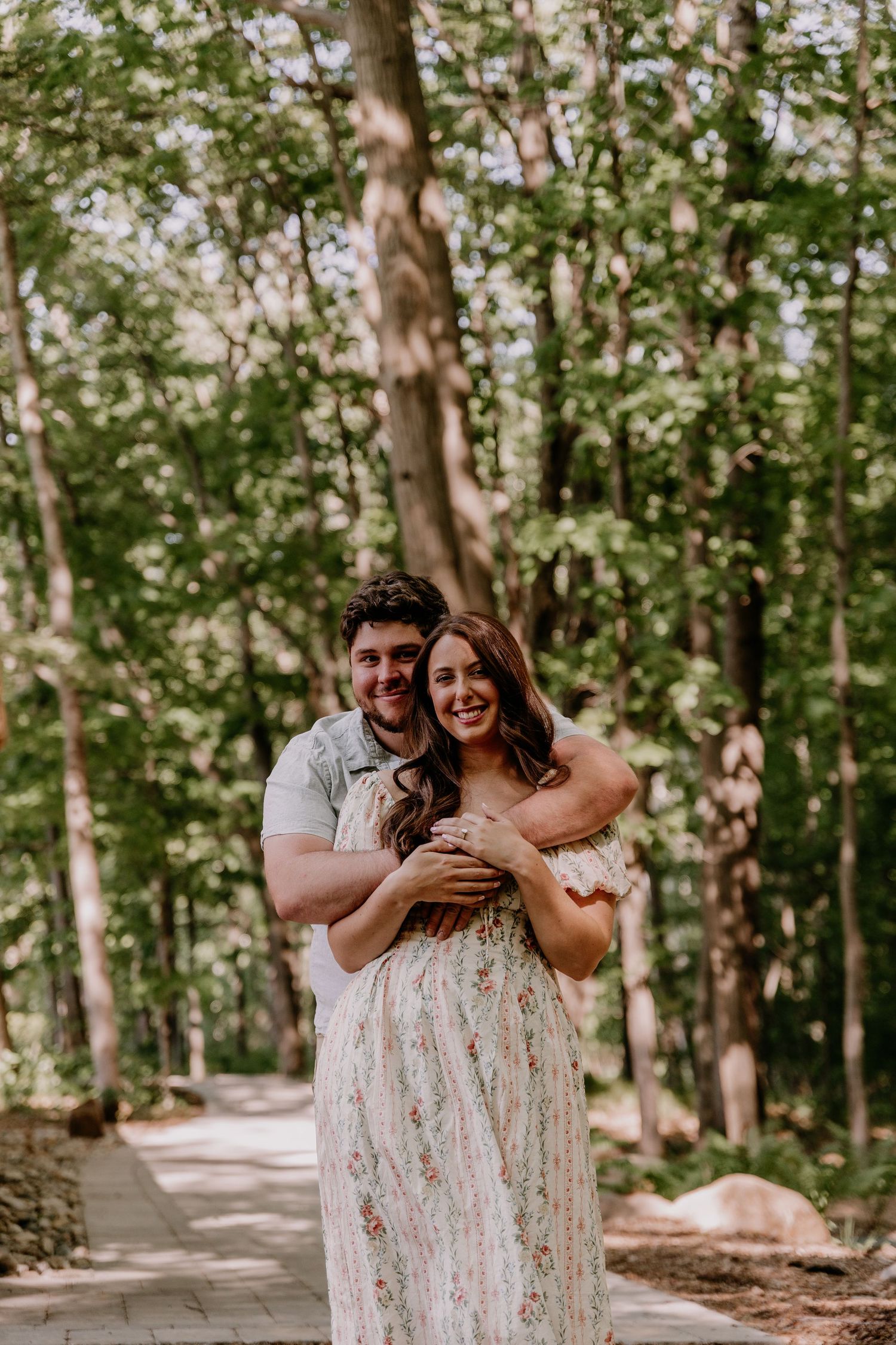A couple embraces and smiles in a lush forest setting, woman in floral maxi dress, man in casual shirt.
