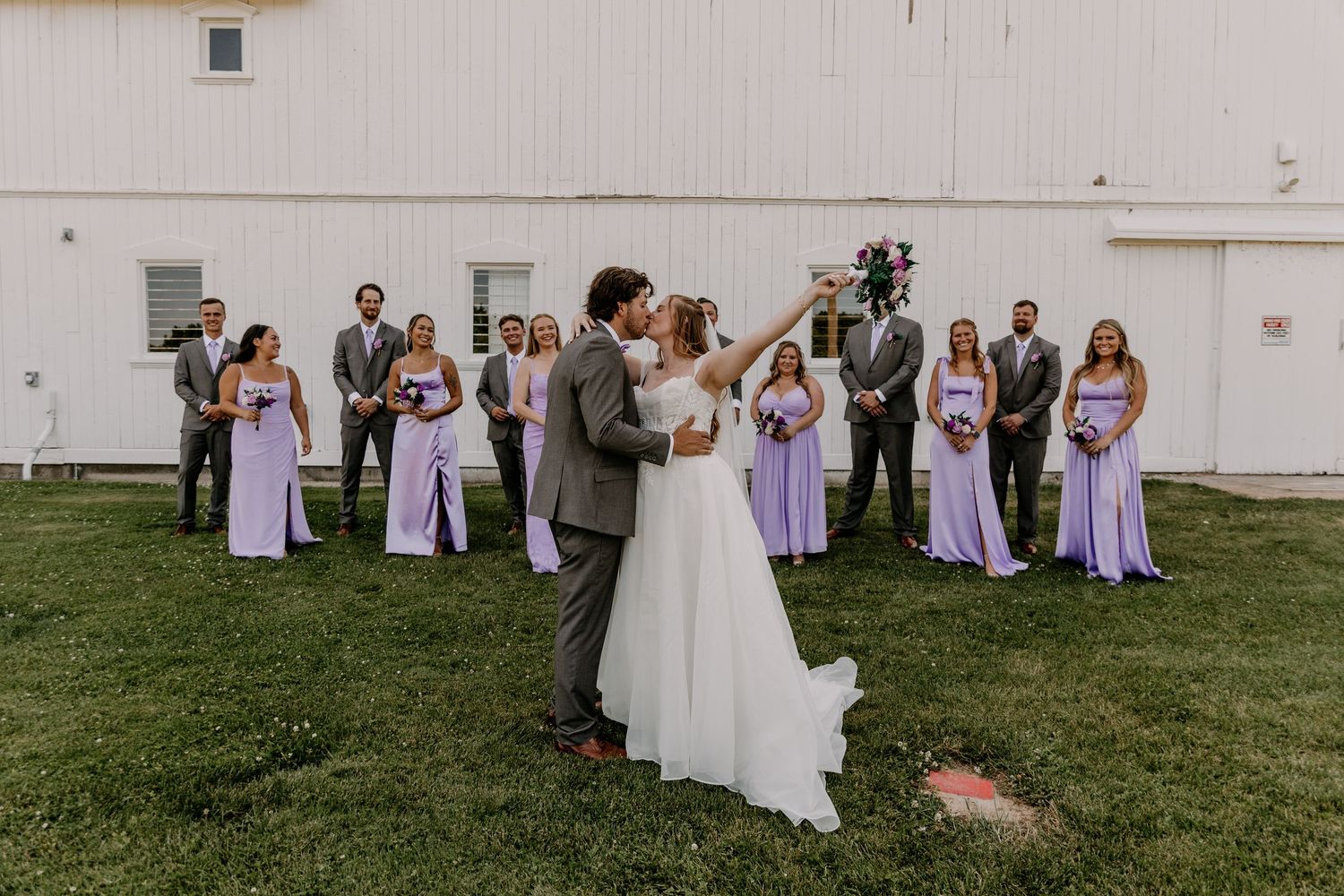 Bride and groom share a kiss while wedding party in lavender and gray stands behind.