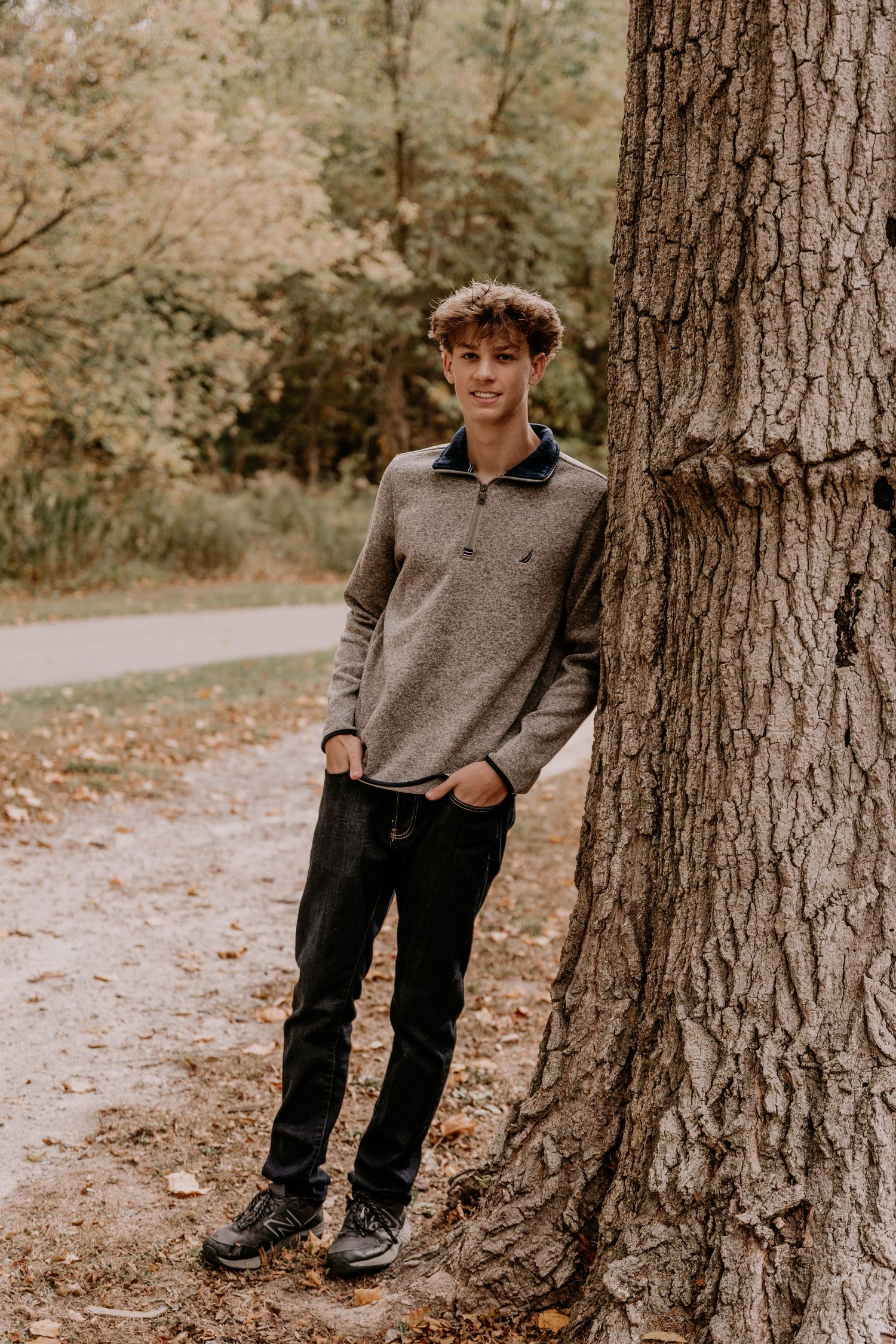 A person in a gray sweater and dark jeans stands casually leaning against a large tree trunk on a wooded path.