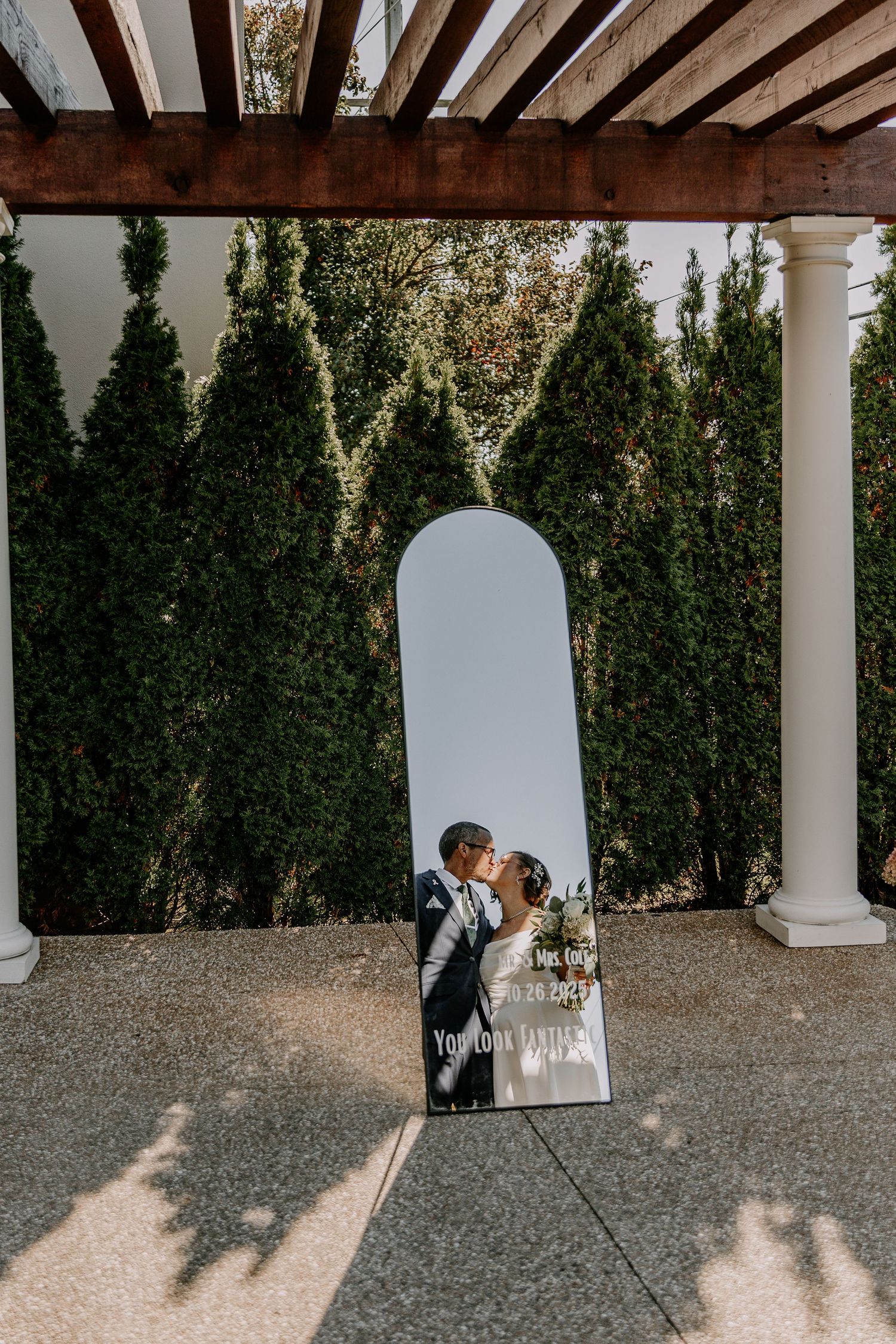 A romantic couple shares a tender moment under a white columned pergola with lush greenery in the background.