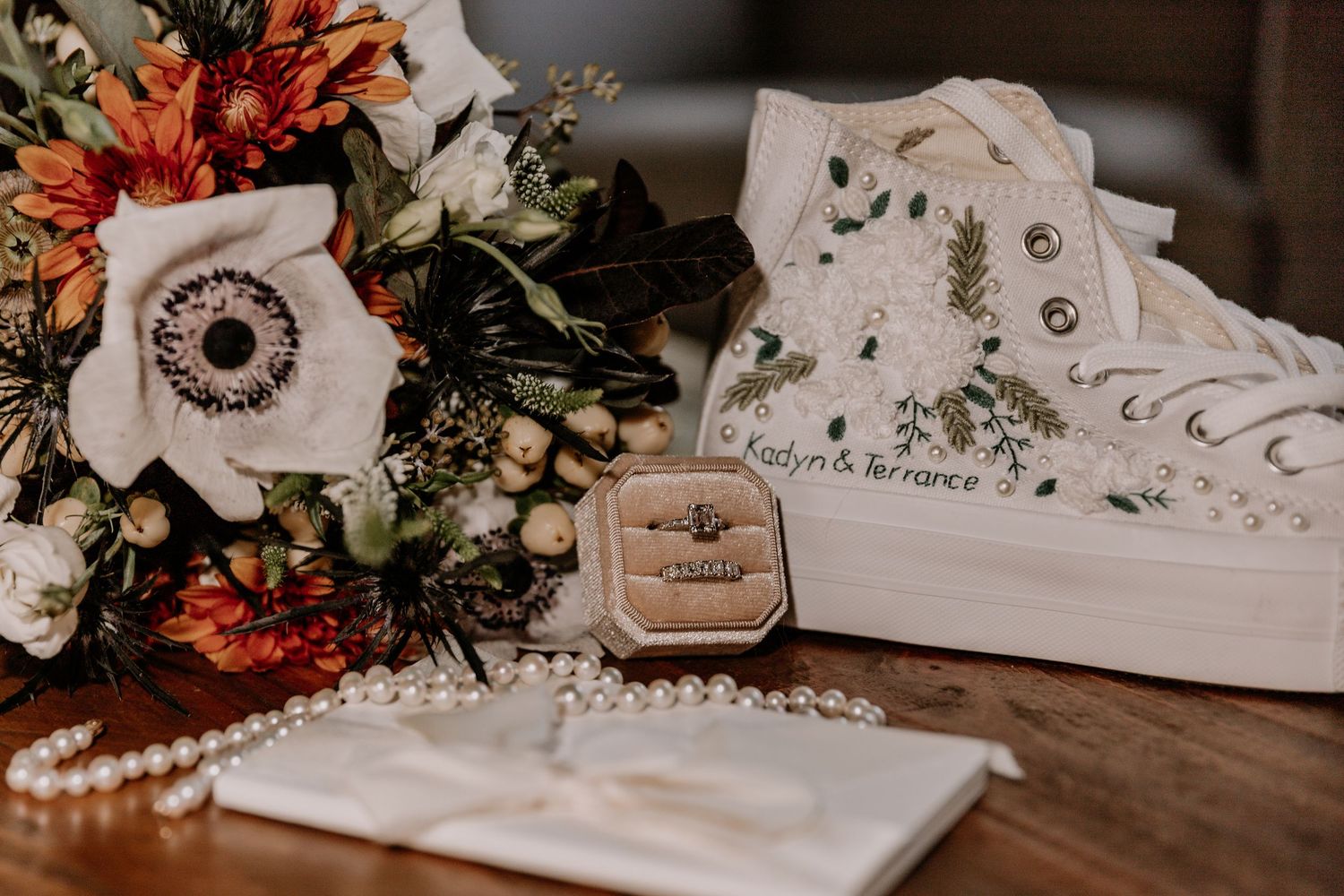 Wedding accessories including leather shoes, engraved invitations, pearls and floral decorations arranged on dark wooden surface.