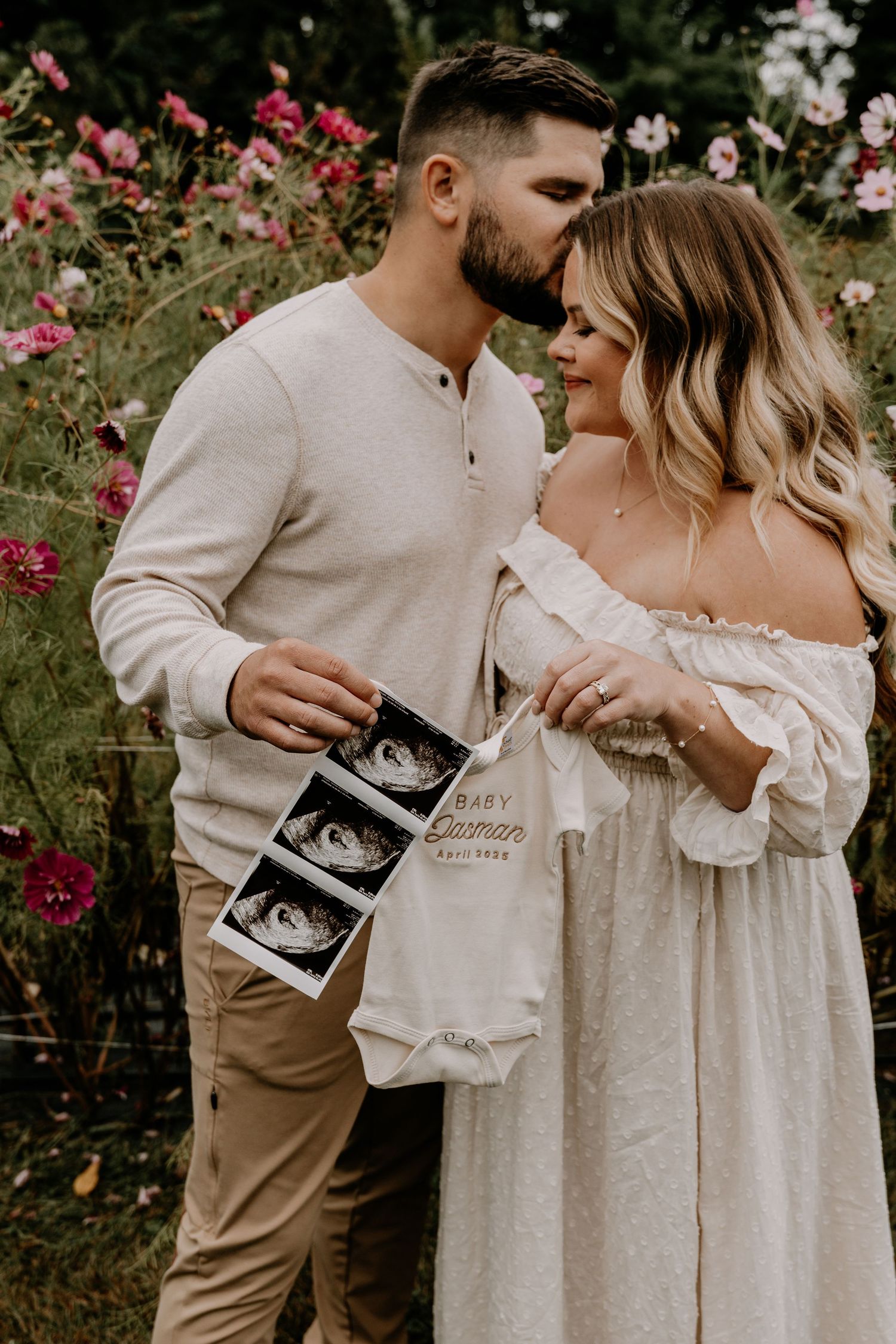 Couple shares an intimate moment in a garden while holding ultrasound photos surrounded by pink flowers.
