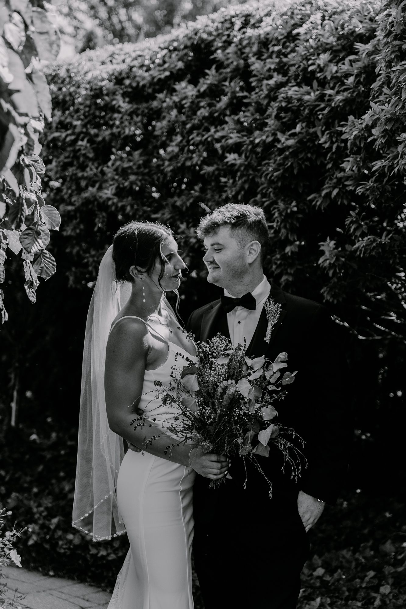 Black and white photo of couple sharing intimate moment in garden with bouquet.