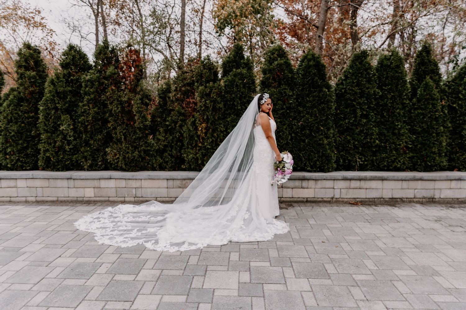 Bride in flowing white dress and cathedral veil poses on stone patio with evergreen trees in background.