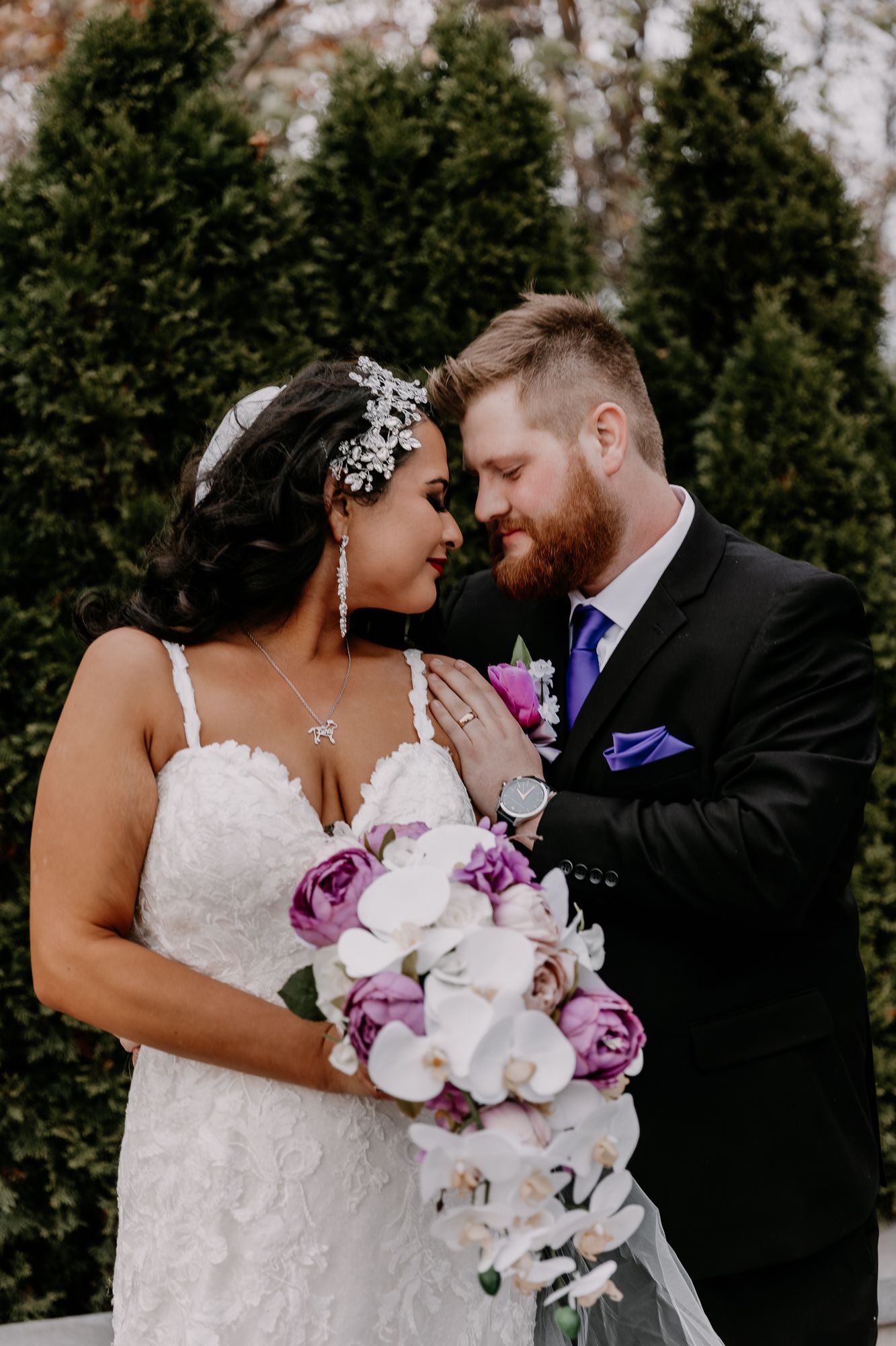 A couple shares a tender moment while holding a purple and white wedding bouquet.