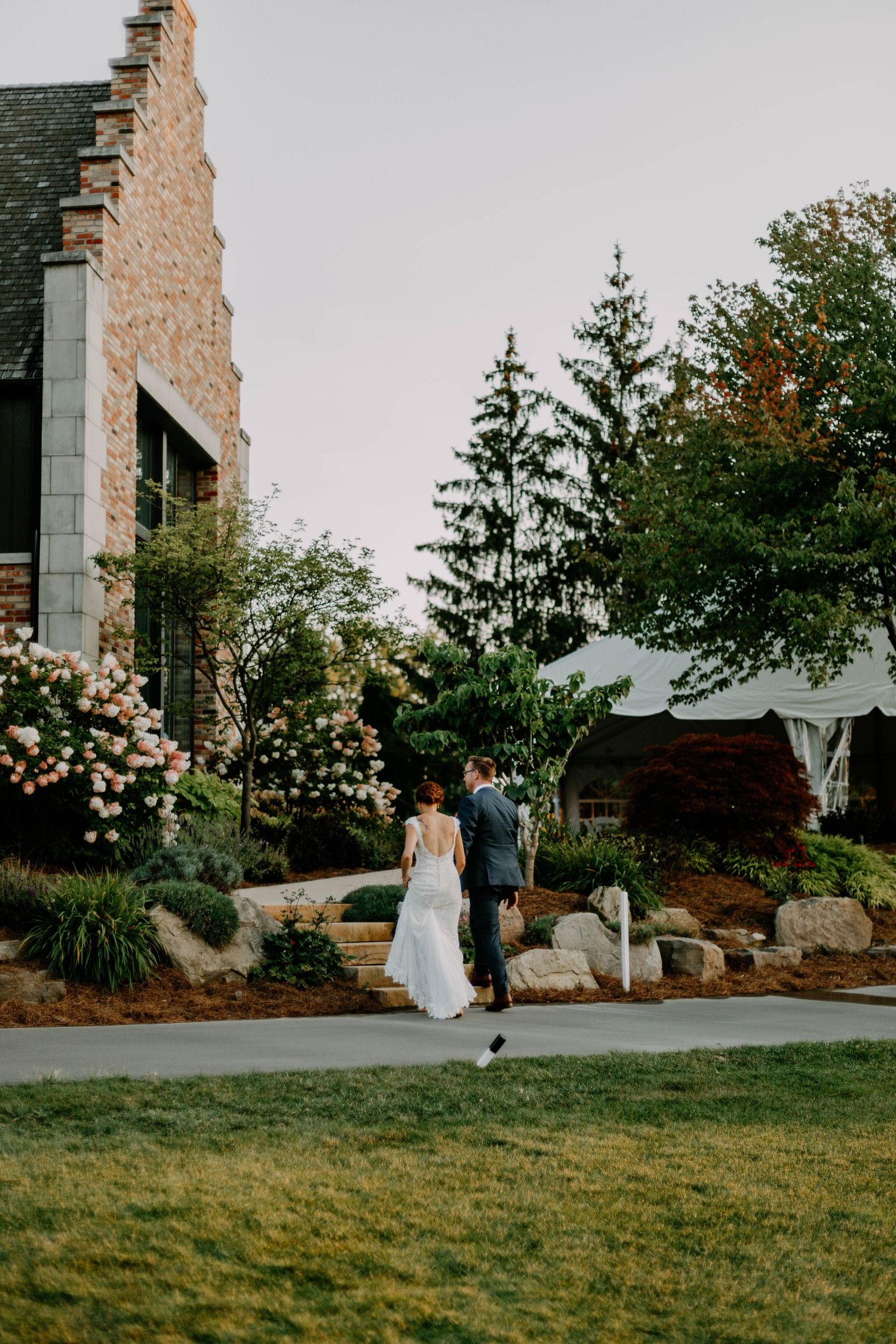 A wedding couple stands outside a stone building surrounded by blooming flowers and landscaping.