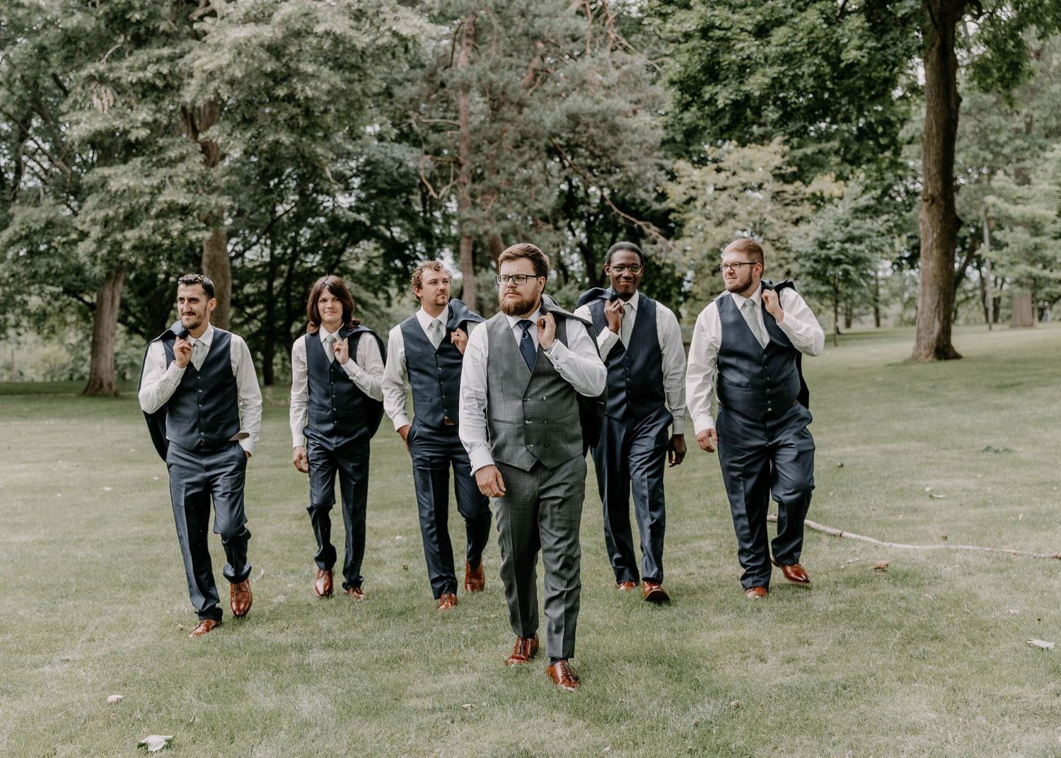 Wedding party walks together across green lawn wearing coordinated gray suits.
